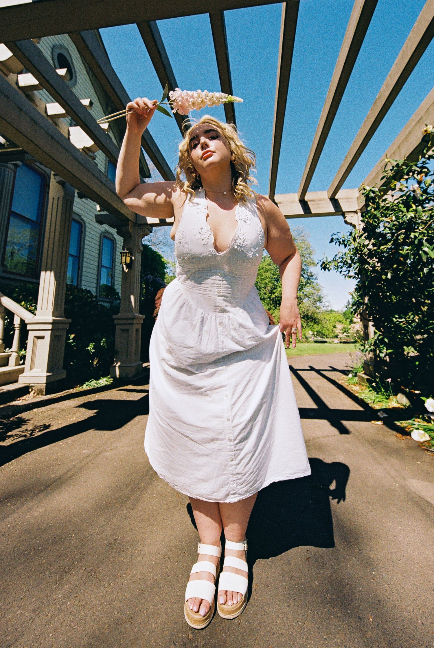 Plus-size woman in white halter sundress holding flower, standing under pergola on sunny day.
