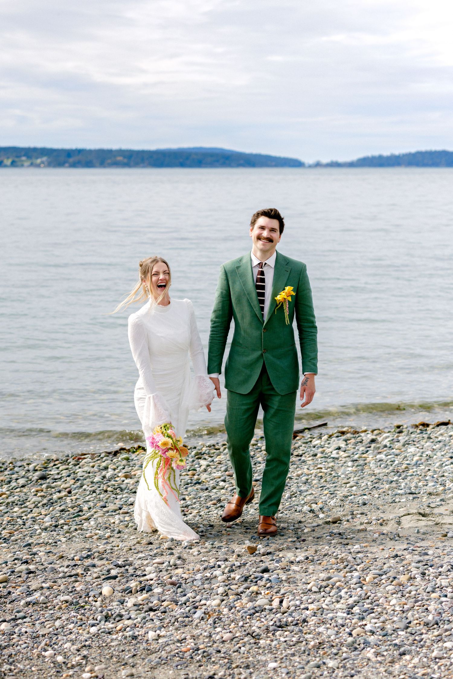 A joyful couple walks on a rocky beach, bride in white dress and groom in green suit holding yellow flowers.