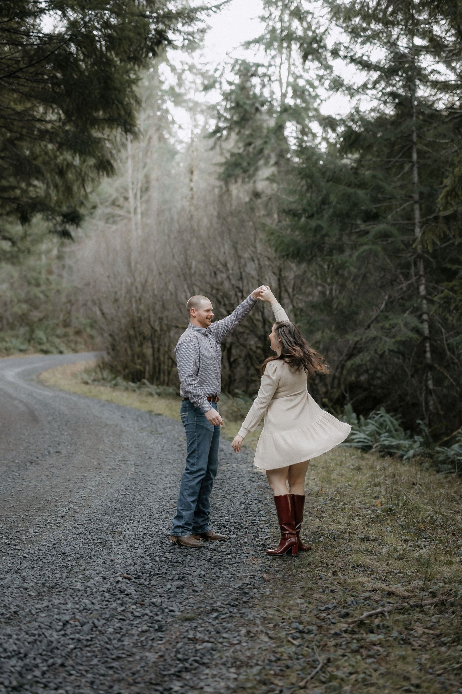 A couple shares playful moments on a gravel path surrounded by evergreen trees in a forest setting.