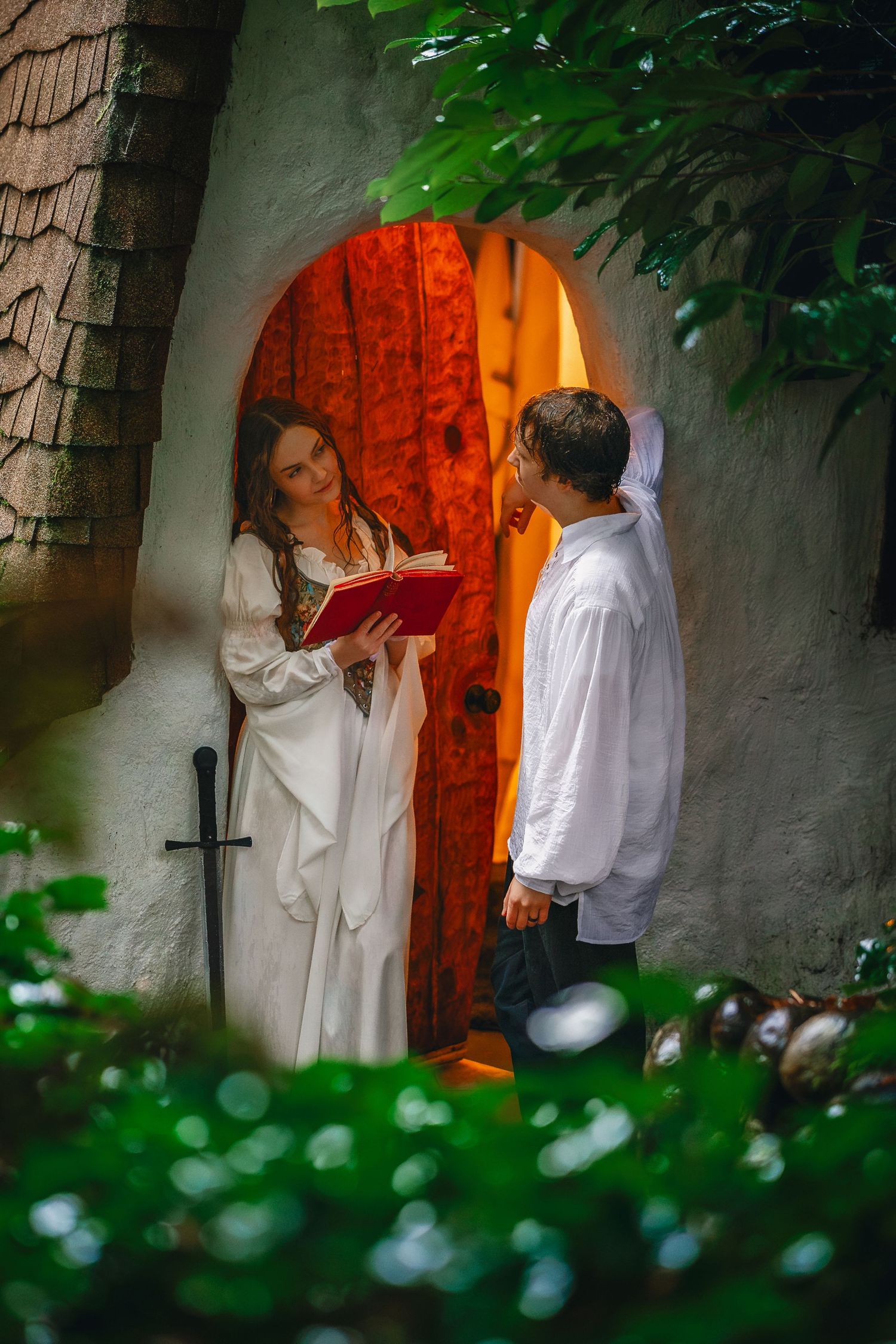 A romantic couple in white attire stands near a rustic wooden door framed by green foliage.