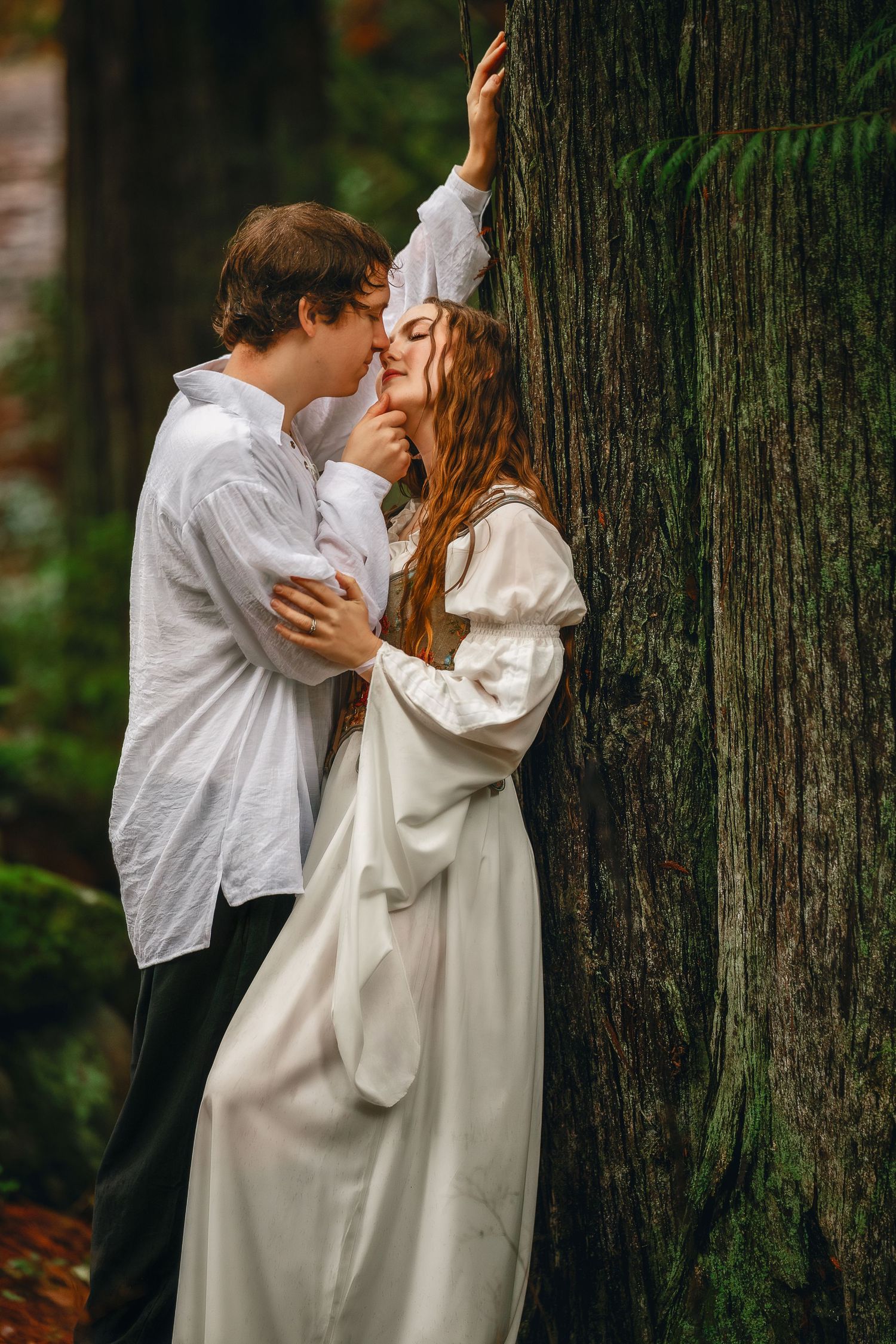 A romantic couple embraces against a large tree trunk in a forest setting wearing white and dark clothing.