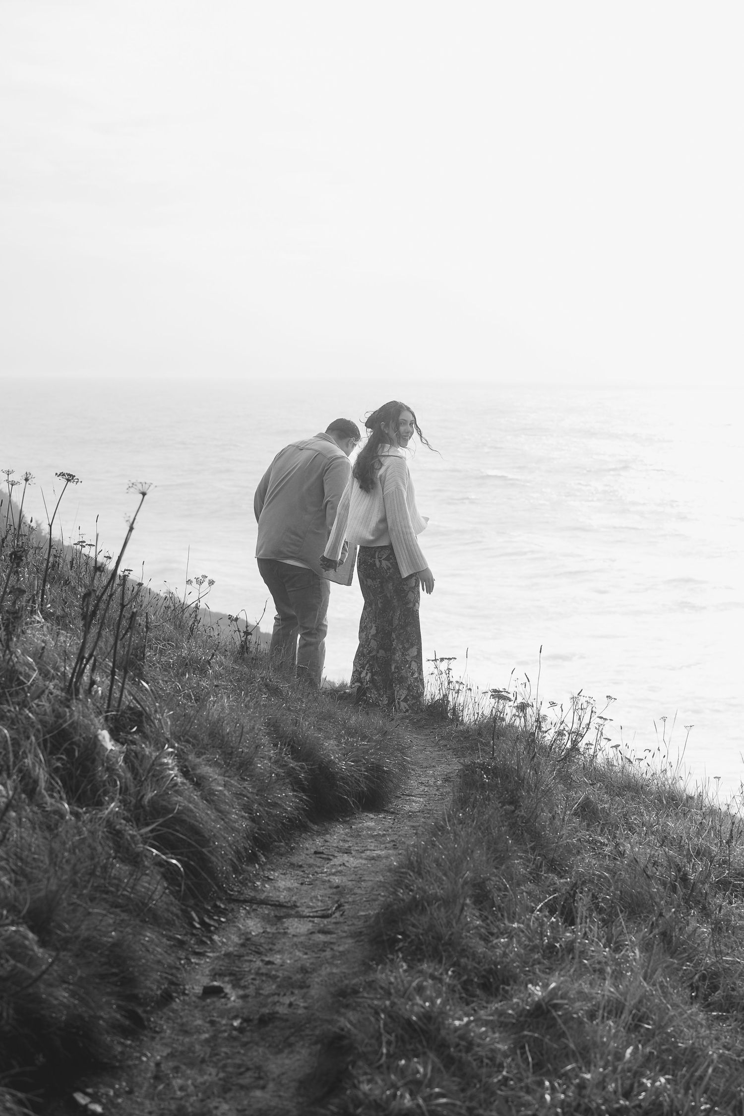 Two figures stand silhouetted against a bright sky on a grassy hillside path overlooking the ocean.