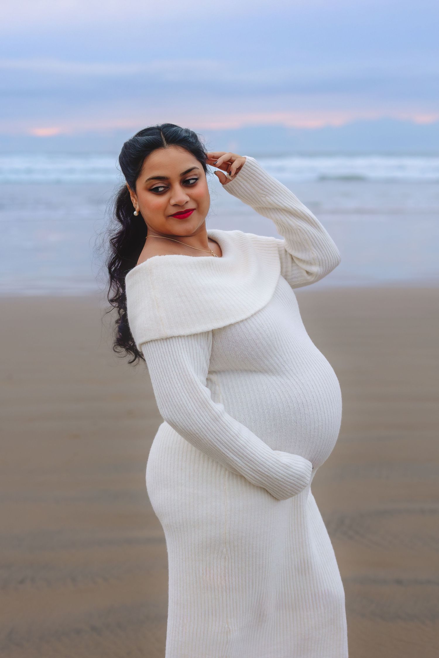 A pregnant woman in a white knit dress poses on a beach at sunset against a pastel sky.