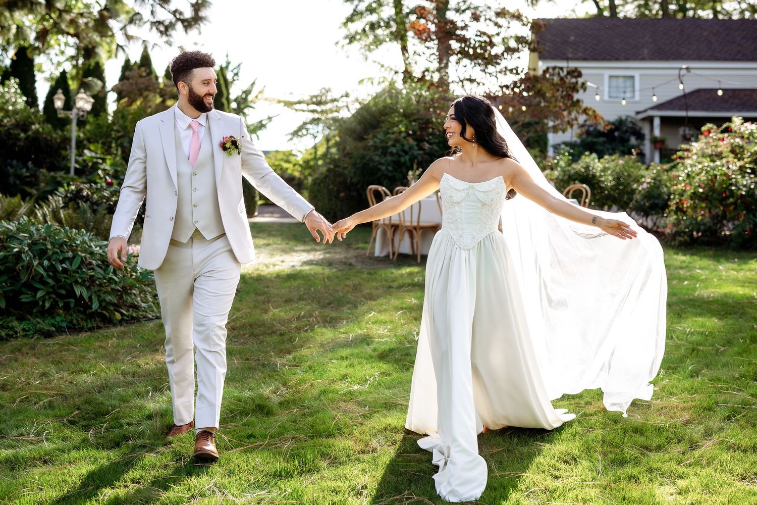 A couple in elegant white wedding attire dances playfully on a sunlit lawn surrounded by greenery.