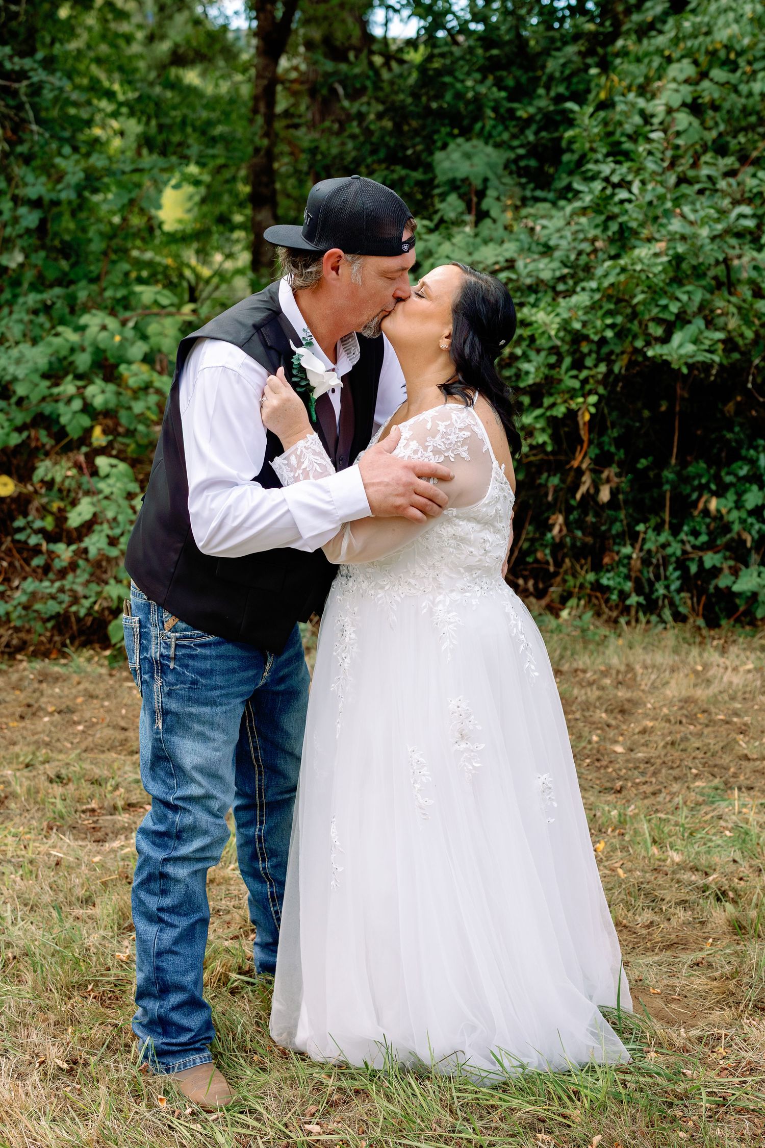 A couple in western wedding attire share romantic kisses in an outdoor wooded setting.
