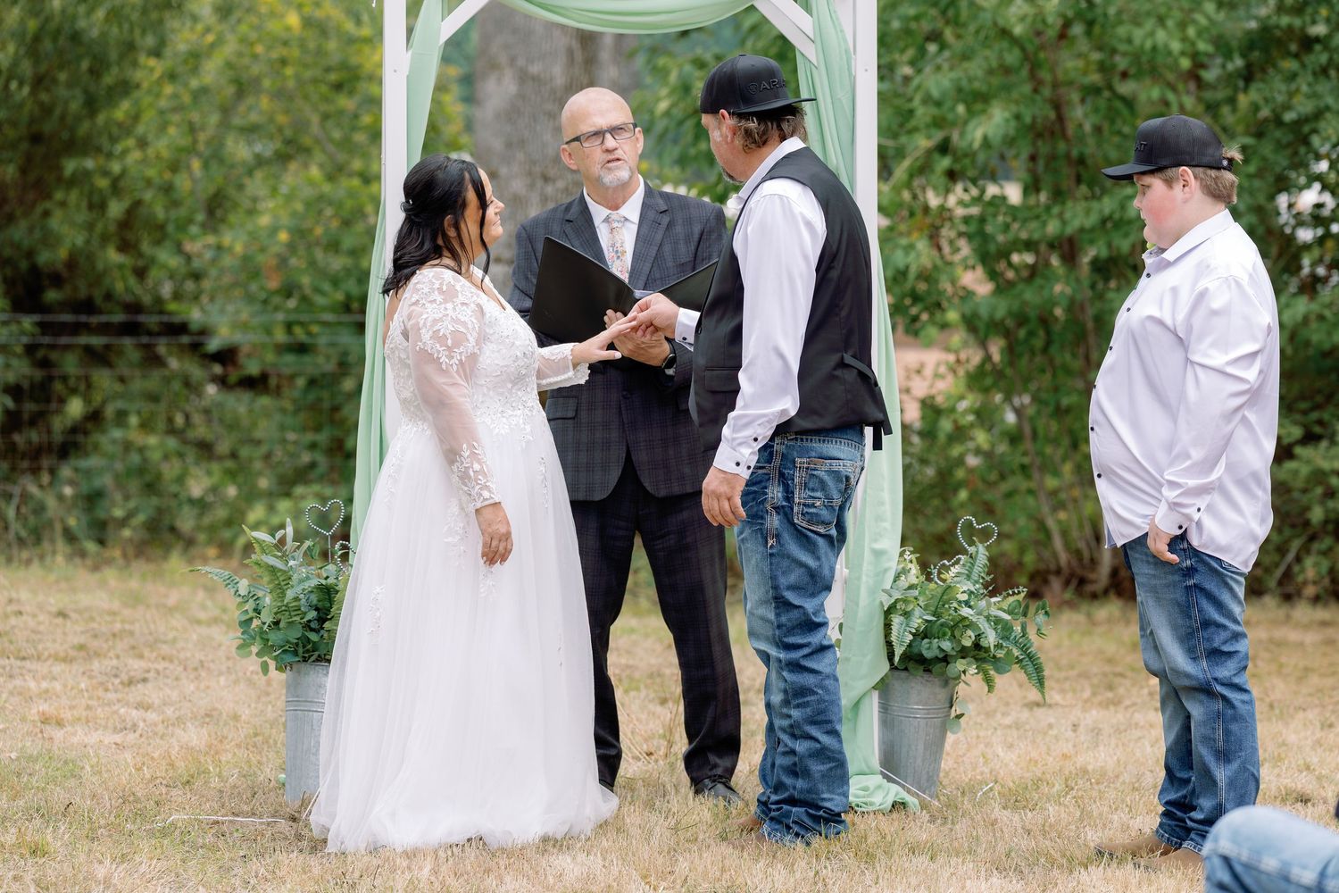 Outdoor wedding ceremony taking place under a white arch with guests gathered in a garden setting.