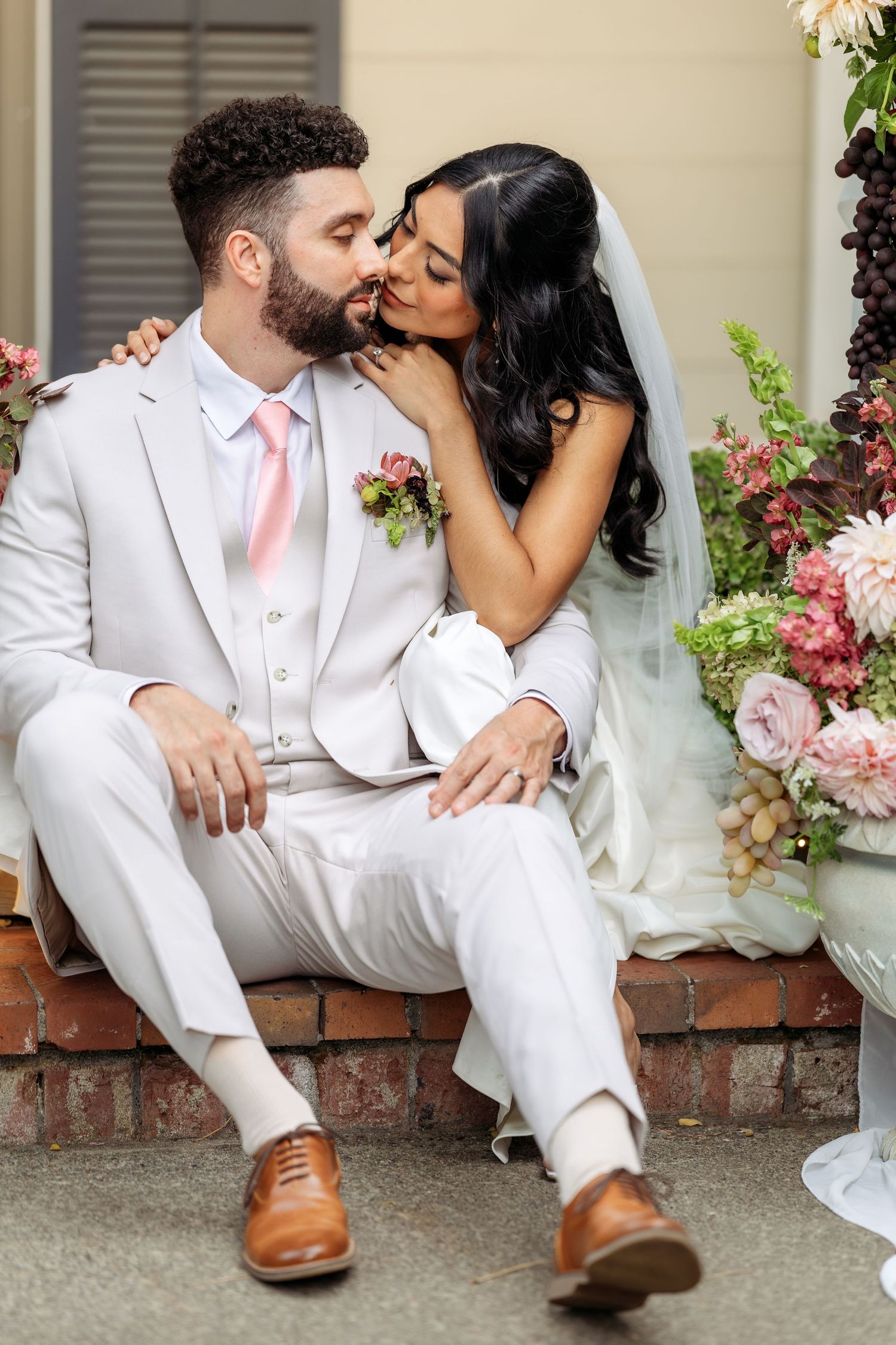 Newlyweds in white attire share intimate moments while sitting on brick steps with floral decorations.