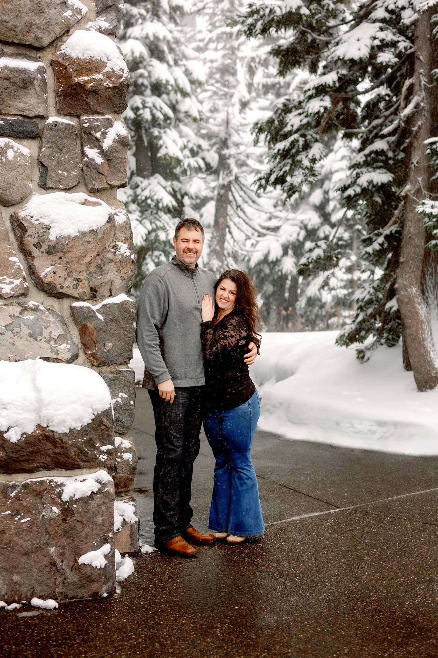 A couple shares a tender moment together on a snowy path surrounded by snow-covered evergreen trees.