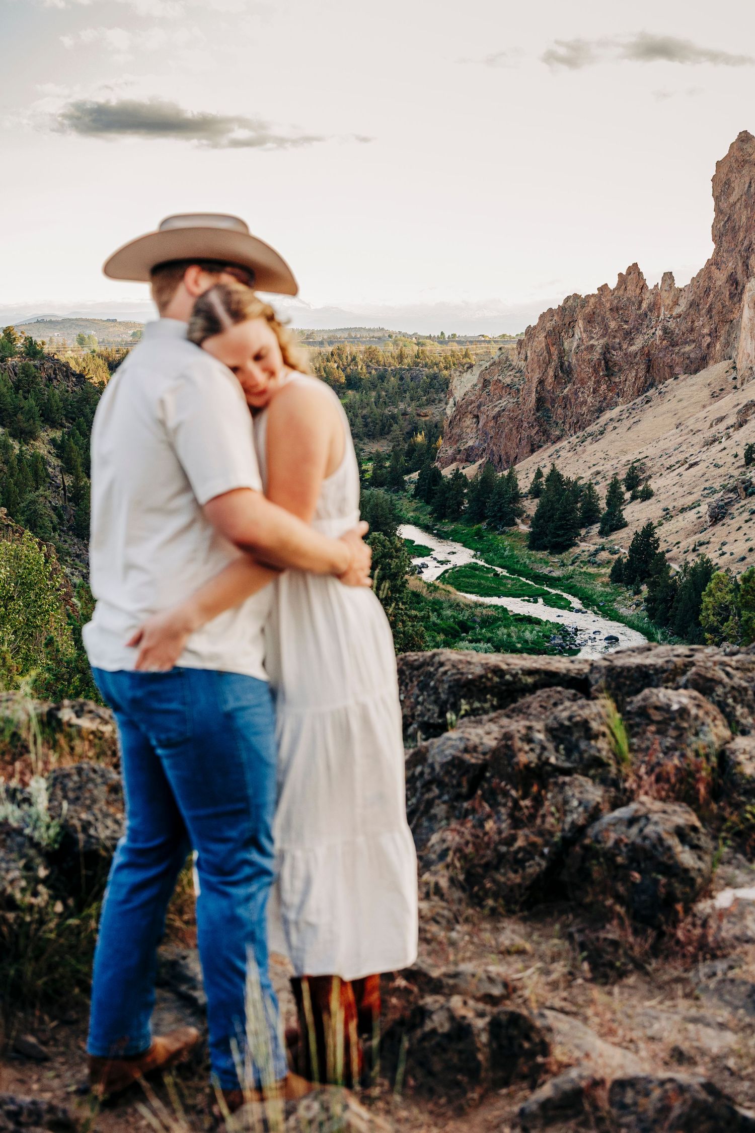 Couple stands together on scenic overlook with dramatic canyon rock formations and winding river below.