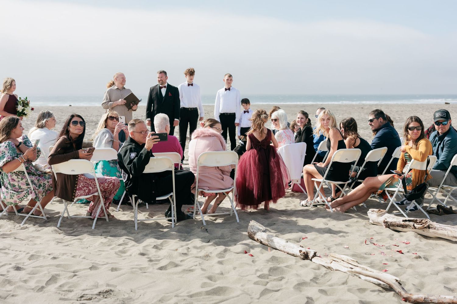 Wedding guests seated in folding chairs on a beach during an outdoor ceremony.