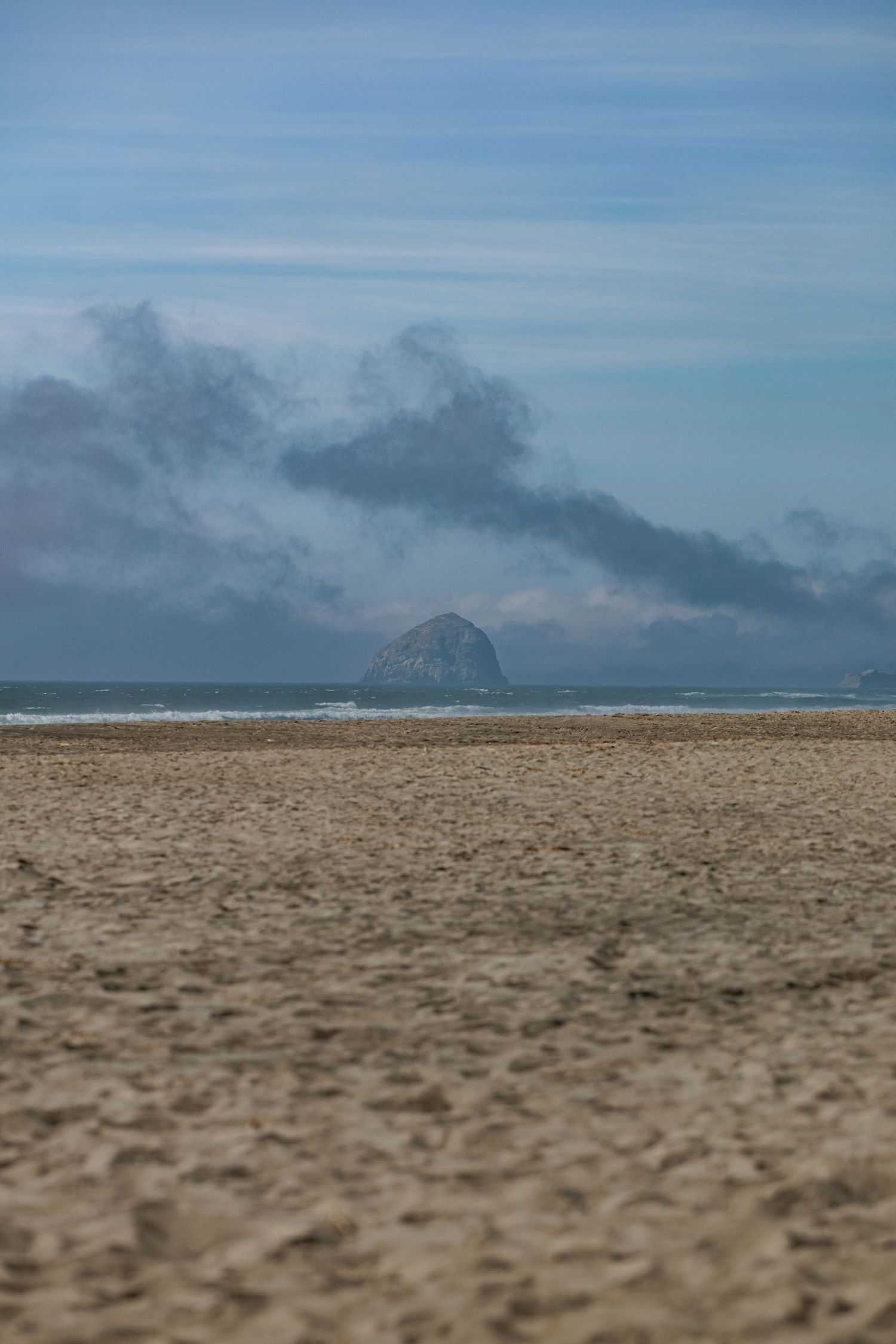 A large rock formation rises from the ocean under dramatic cloudy skies along a sandy beach.
