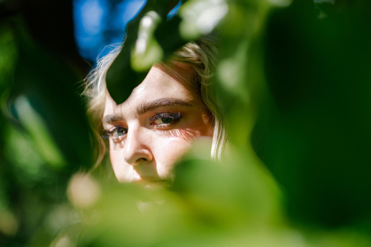 Close-up of a young woman's face partially hidden behind lush green leaves in natural sunlight.