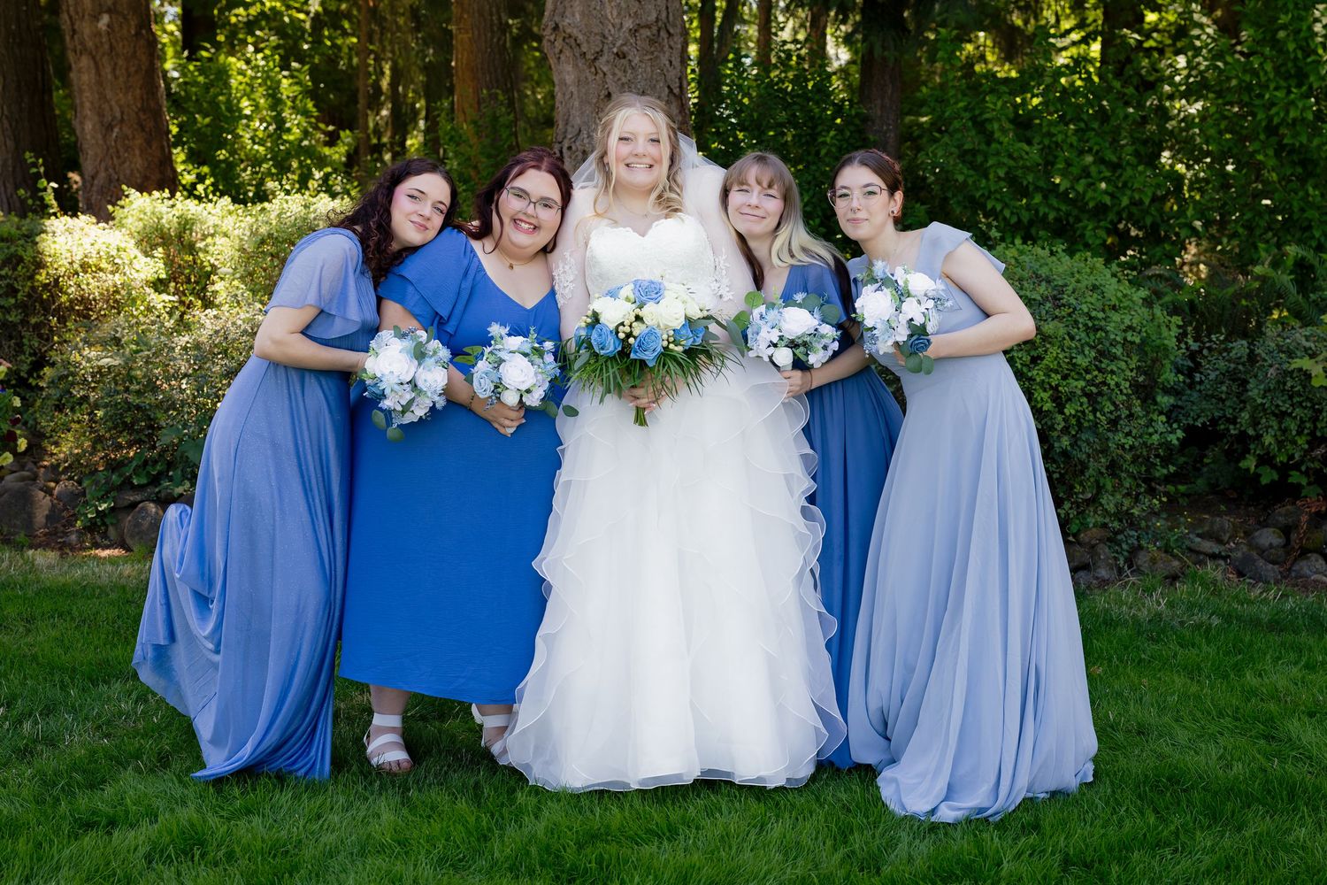 Bride in white gown with four bridesmaids in blue dresses holding bouquets in a garden setting.