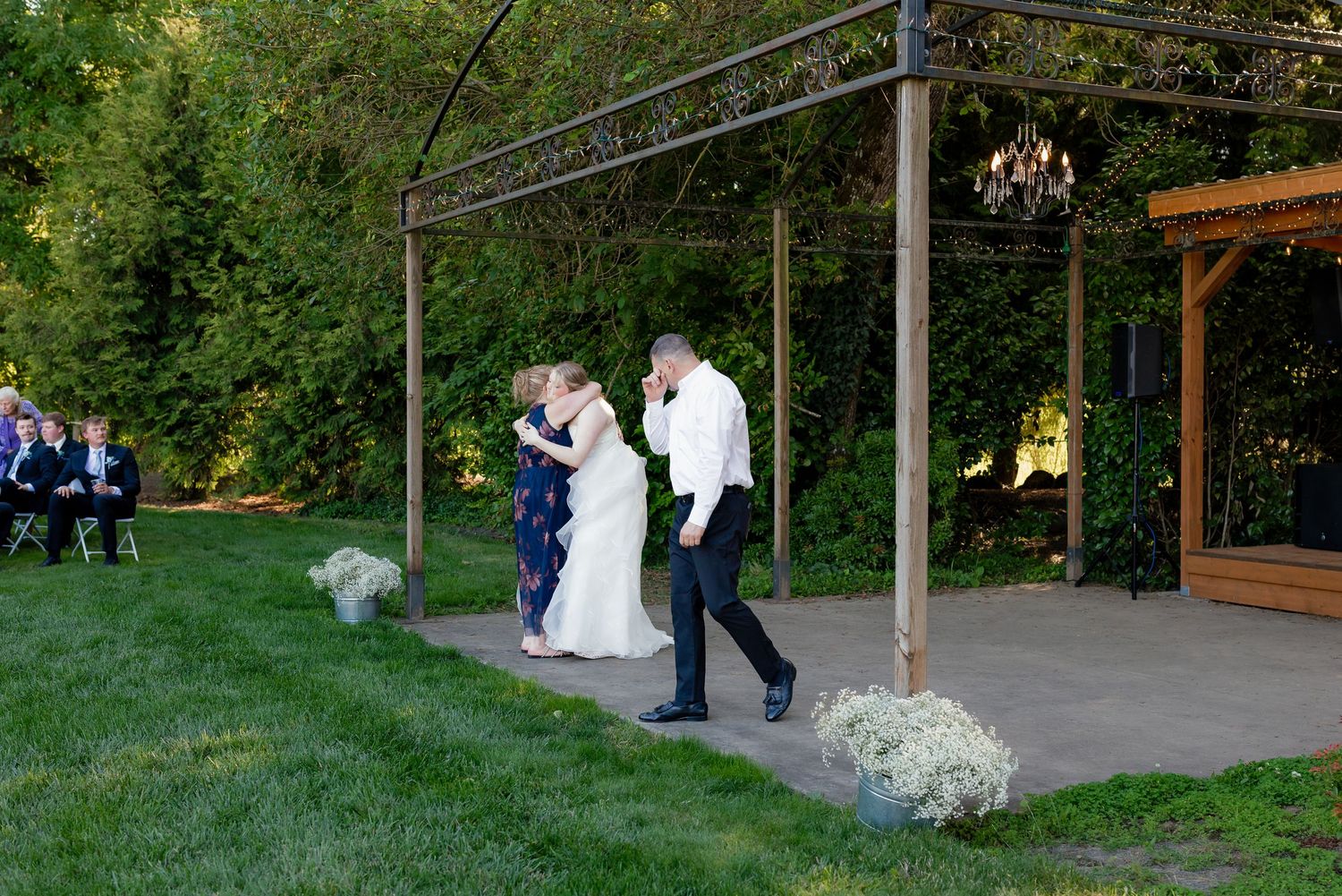 Bride walks down the aisle with escort at an outdoor wedding ceremony beneath a wooden pergola.