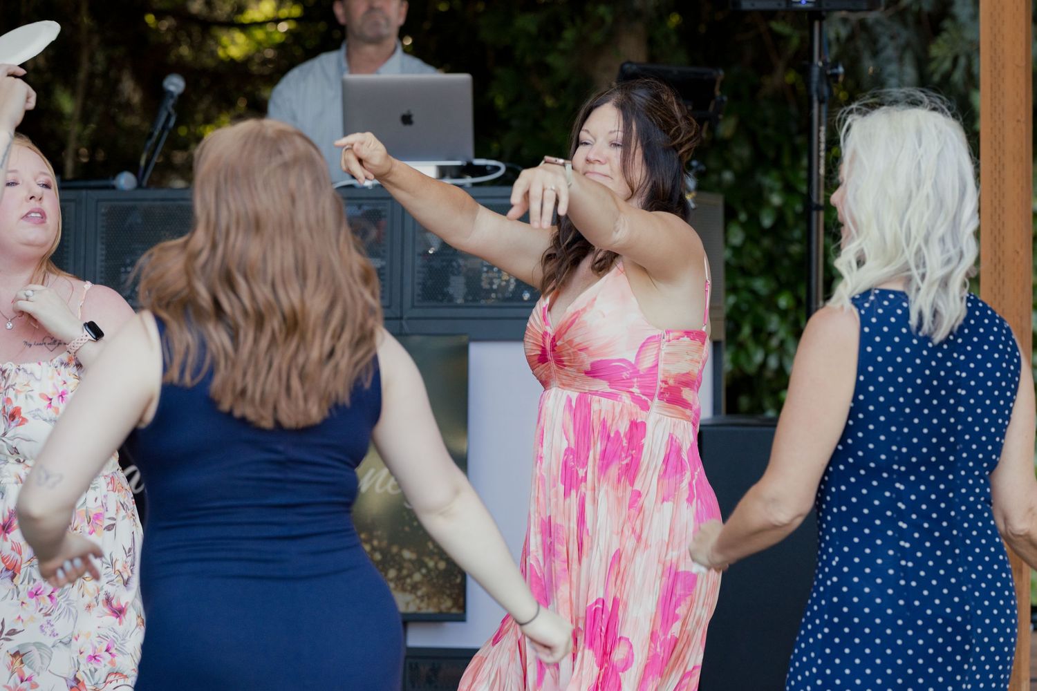 Four women in colorful dresses dance together at outdoor wedding reception with DJ visible in background.