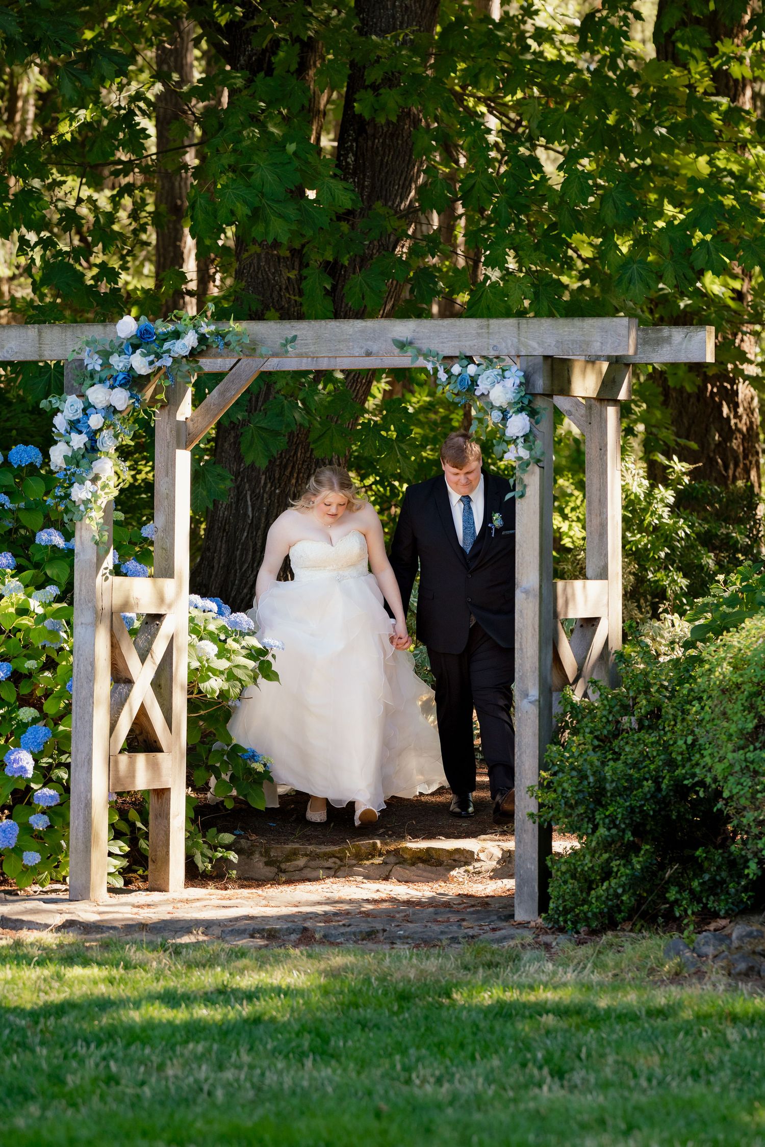 Bride in white gown and groom in black suit walk hand-in-hand through rustic wooden arch with blue hydrangeas.