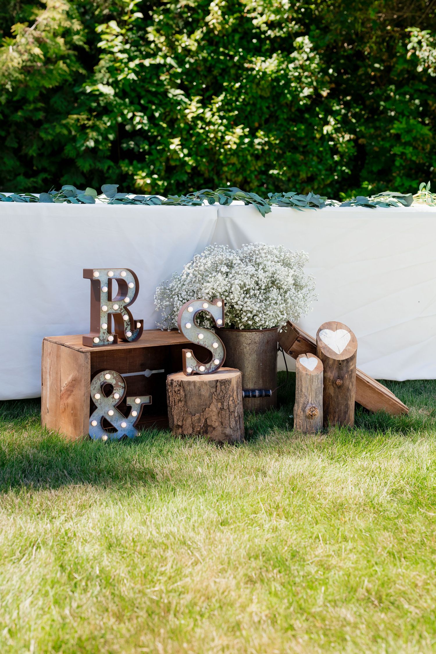 Rustic wedding decor with marquee letters R, S, ampersand on wood stumps and crates with white floral arrangement outdoors.