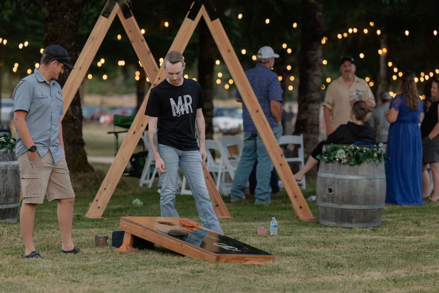 Wedding guest plays cornhole on grass at evening outdoor reception with string lights and wooden A-frame decor.