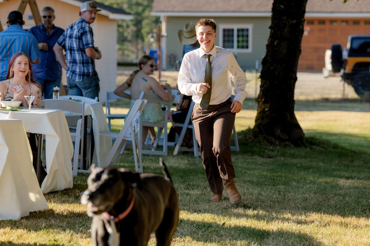 A young man in a tie runs across a sunny outdoor wedding reception lawn near a black dog.