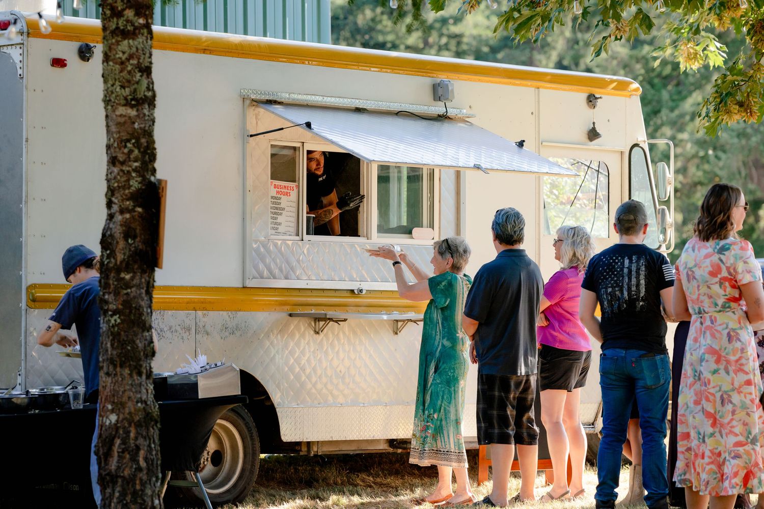 Wedding guests line up at a white food truck parked under trees at an outdoor reception venue.