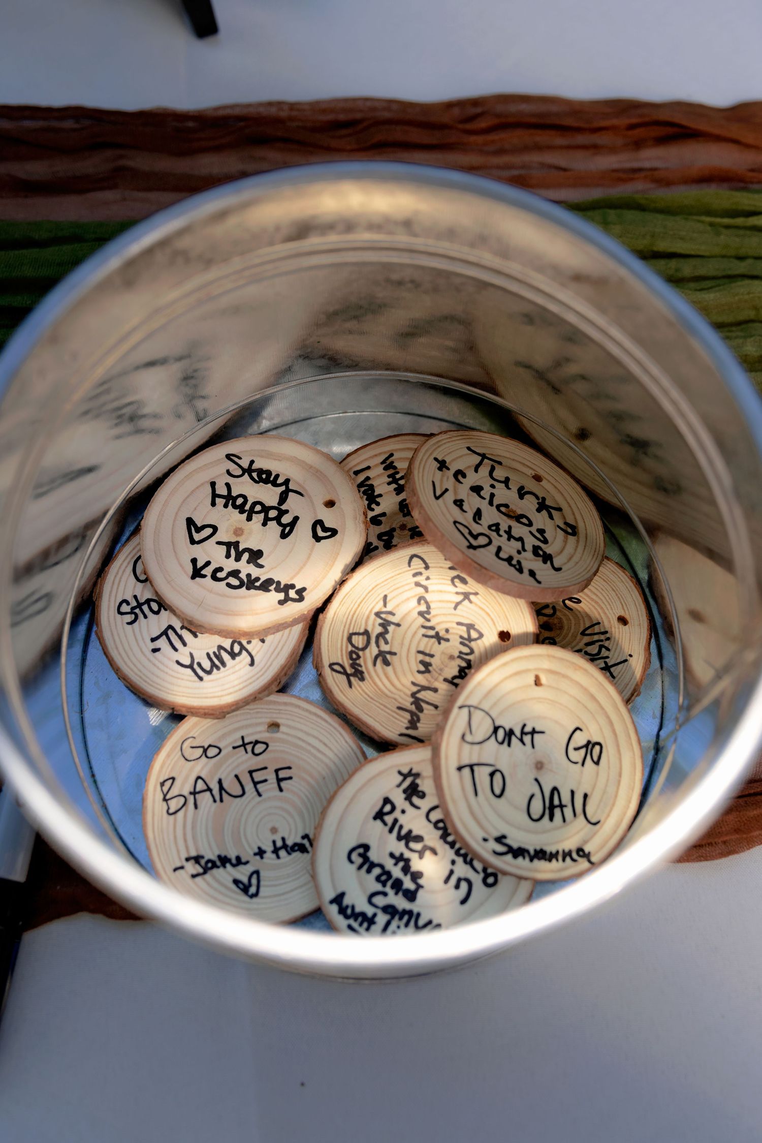 Wooden disc wedding guest wishes in a silver bowl, featuring handwritten messages including Go to Banff and Don't Go to Jail.