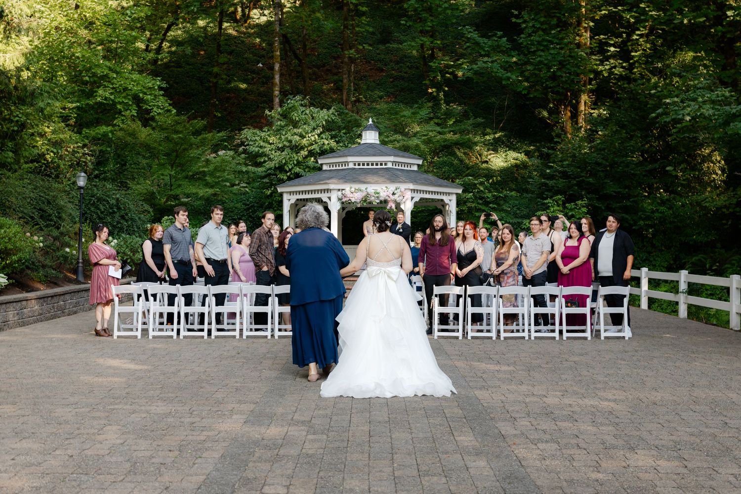 Outdoor wedding ceremony at a gazebo surrounded by lush green trees, with guests standing as the bride and groom walk down the aisle.