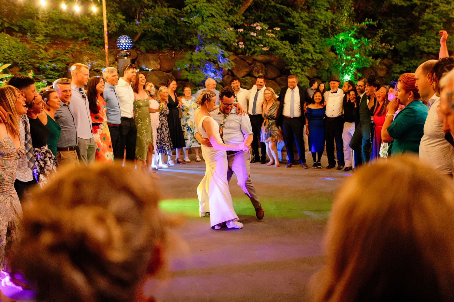 Couple shares first dance at outdoor wedding reception surrounded by guests under string lights at night.