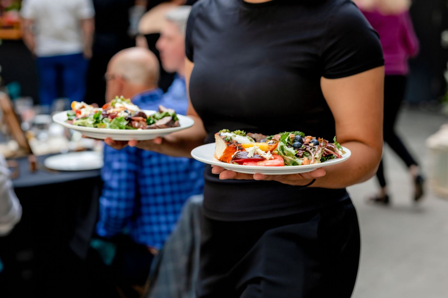 Waitress carrying two plates of fresh gourmet salads at an outdoor catering event.
