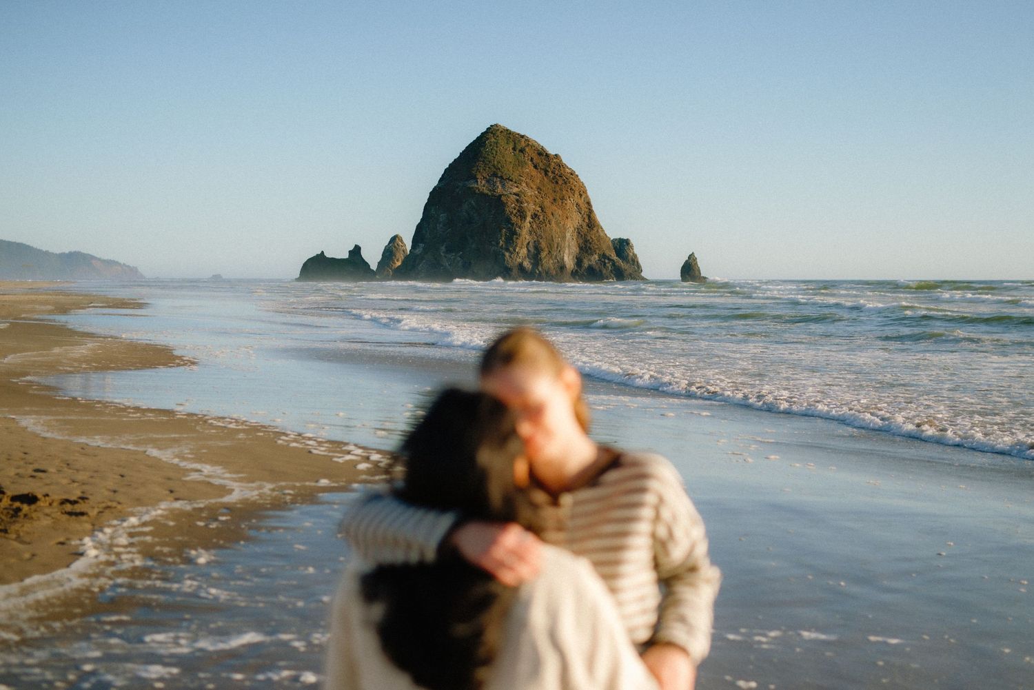 Couple embracing on Cannon Beach with iconic Haystack Rock in the background on a clear sunny day.