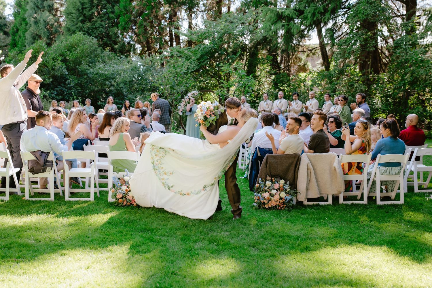 Groom lifting bride during outdoor wedding ceremony on green lawn with guests seated in white chairs among lush trees.