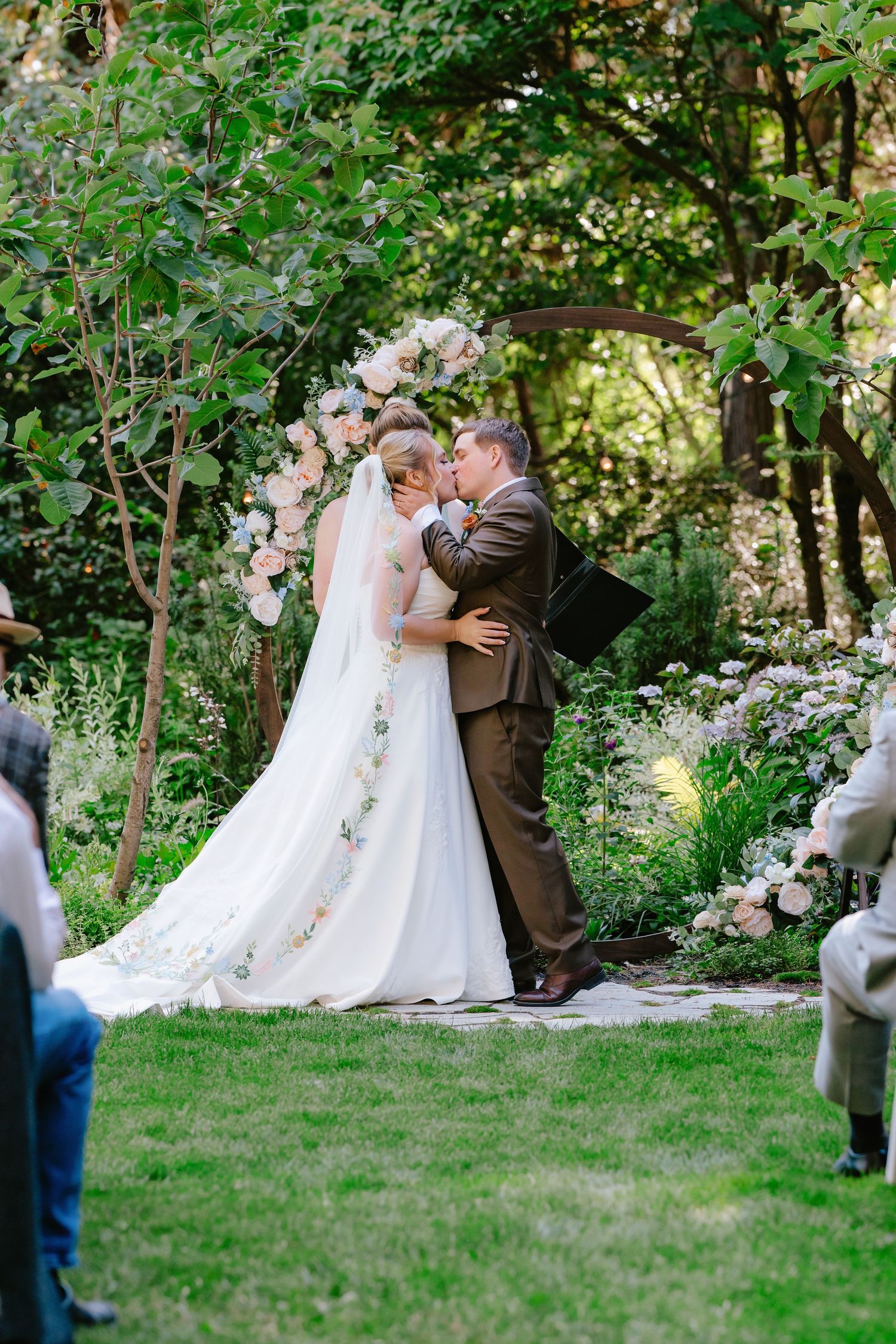 Bride and groom share first kiss under floral arch at lush garden wedding ceremony.