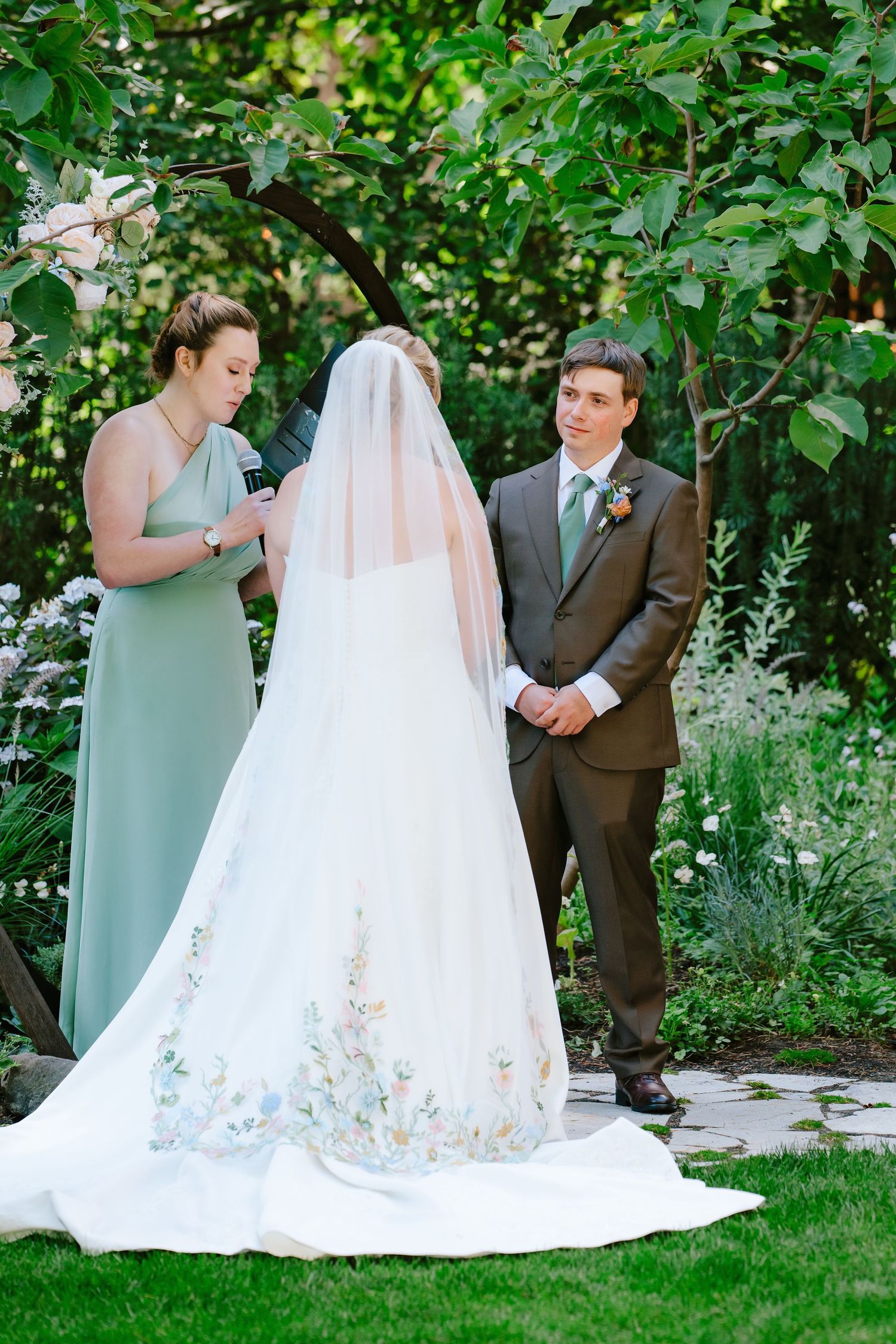 Bride in embroidered veil stands with groom in brown suit during outdoor garden wedding ceremony.