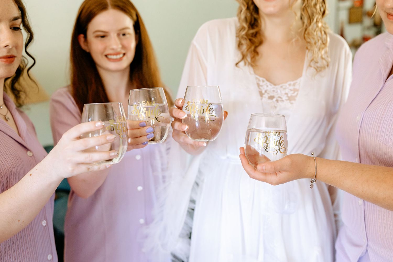 Bridesmaids in pink robes toasting with stemless wine glasses alongside bride in white lace robe.