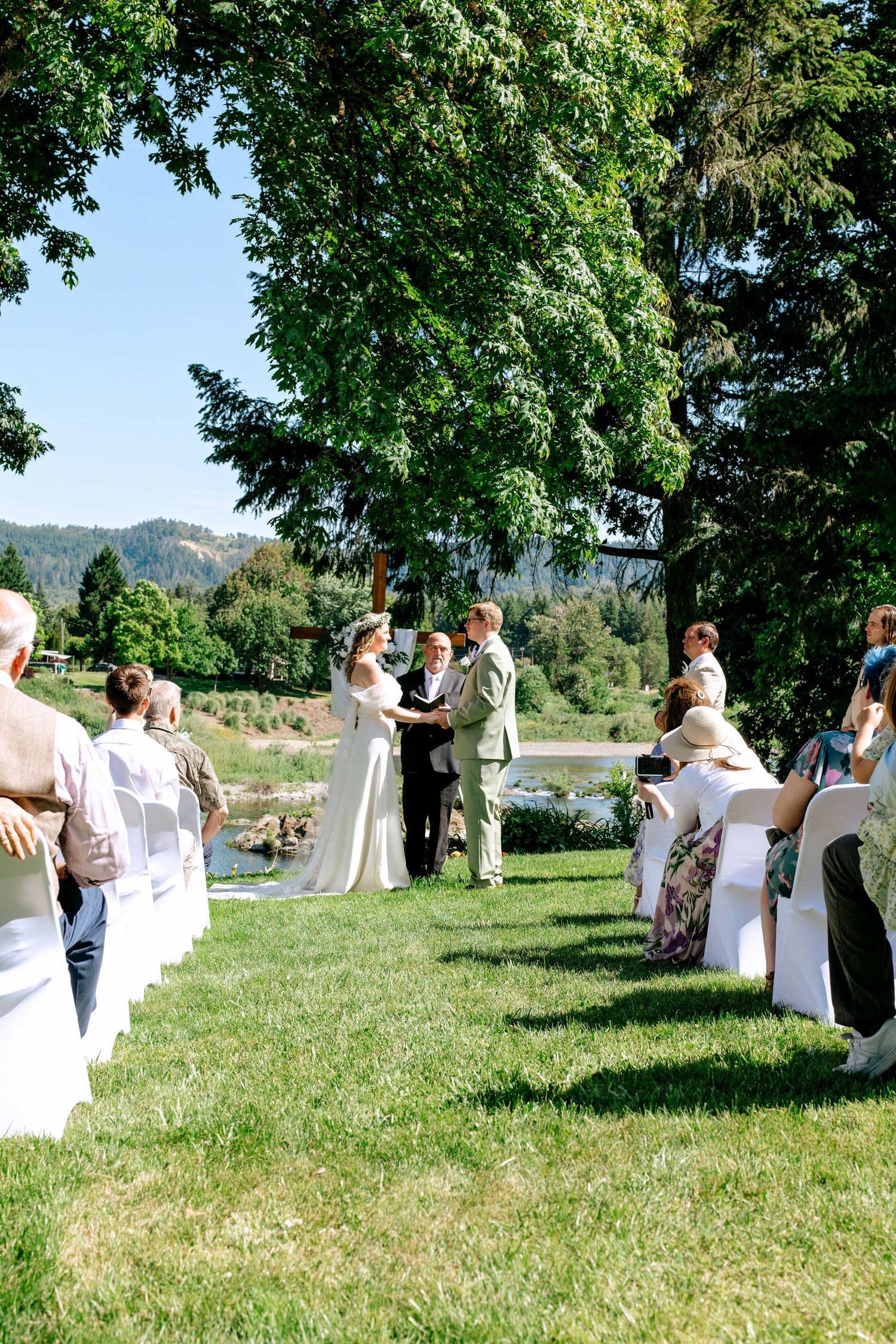Outdoor wedding ceremony with bride and groom exchanging vows on green lawn surrounded by tall trees and mountain backdrop.
