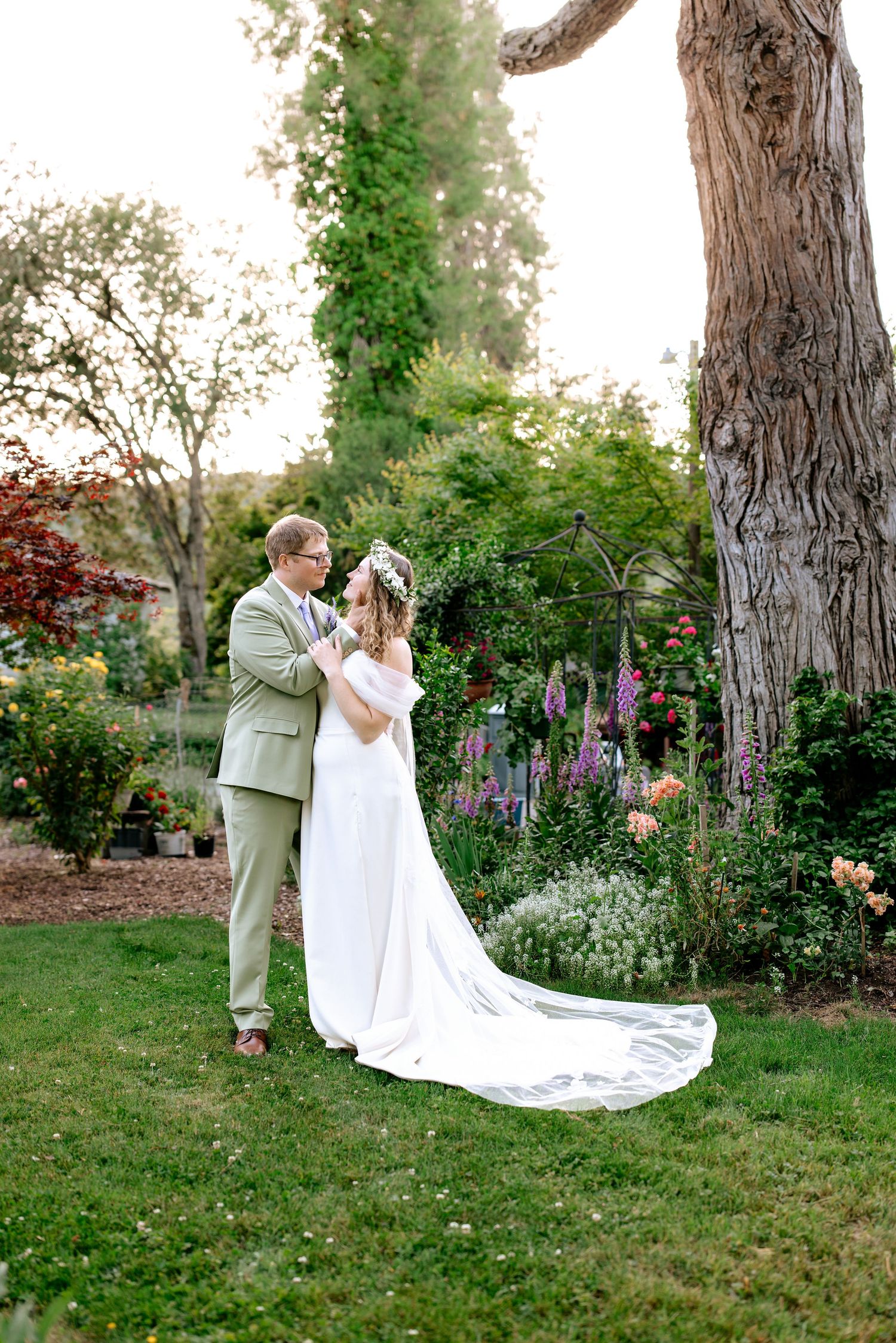 Bride and groom embrace in a lush garden, she wearing a flowing white gown with train and floral crown.