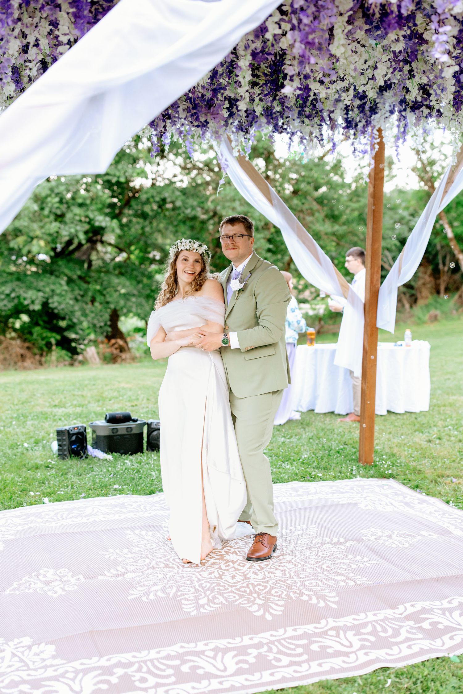 Bride and groom pose together under a beautiful purple wisteria-adorned outdoor wedding canopy.