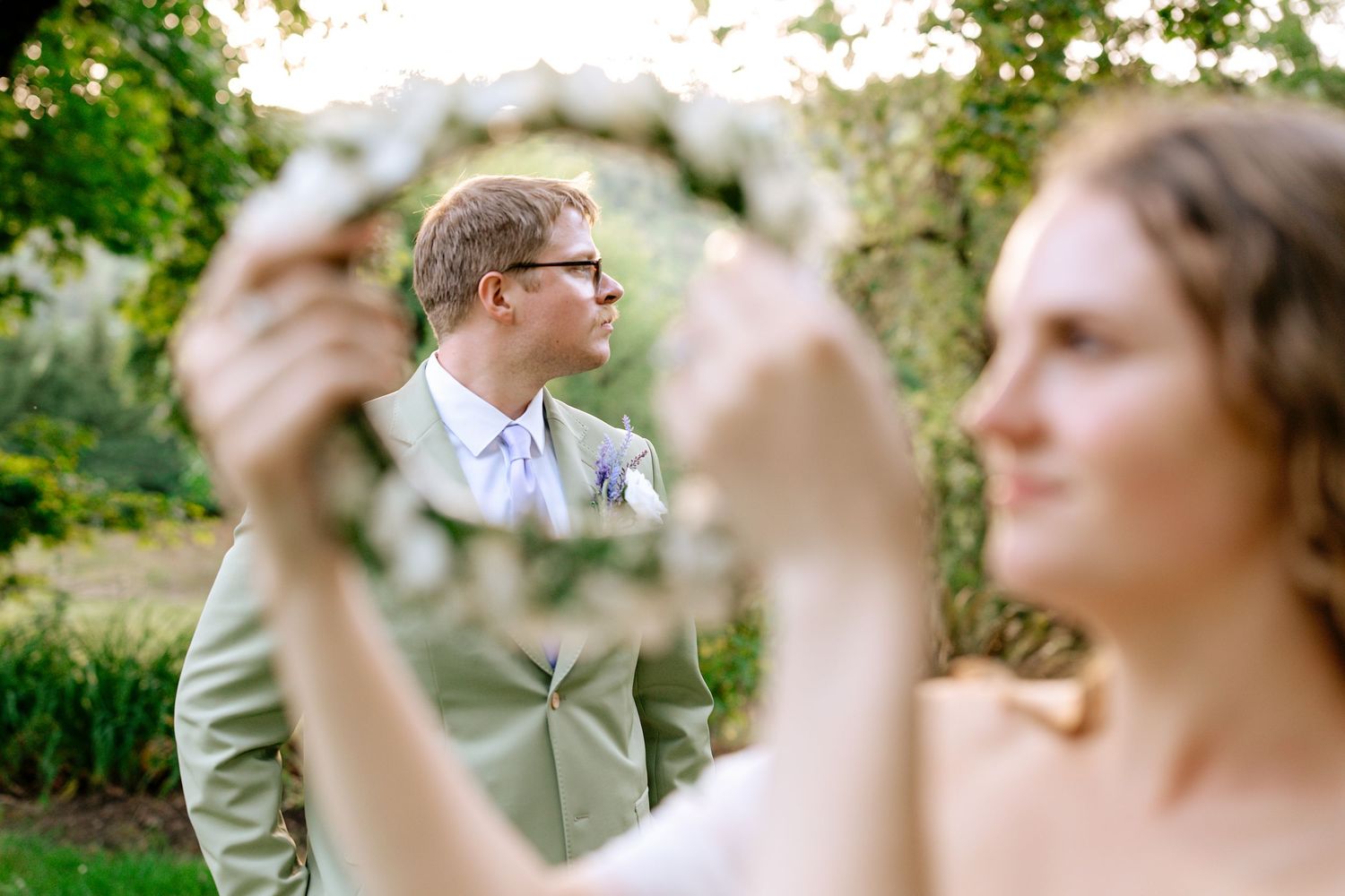 Groom in tan suit reflected in a floral wreath mirror held by bride during creative outdoor wedding portrait.