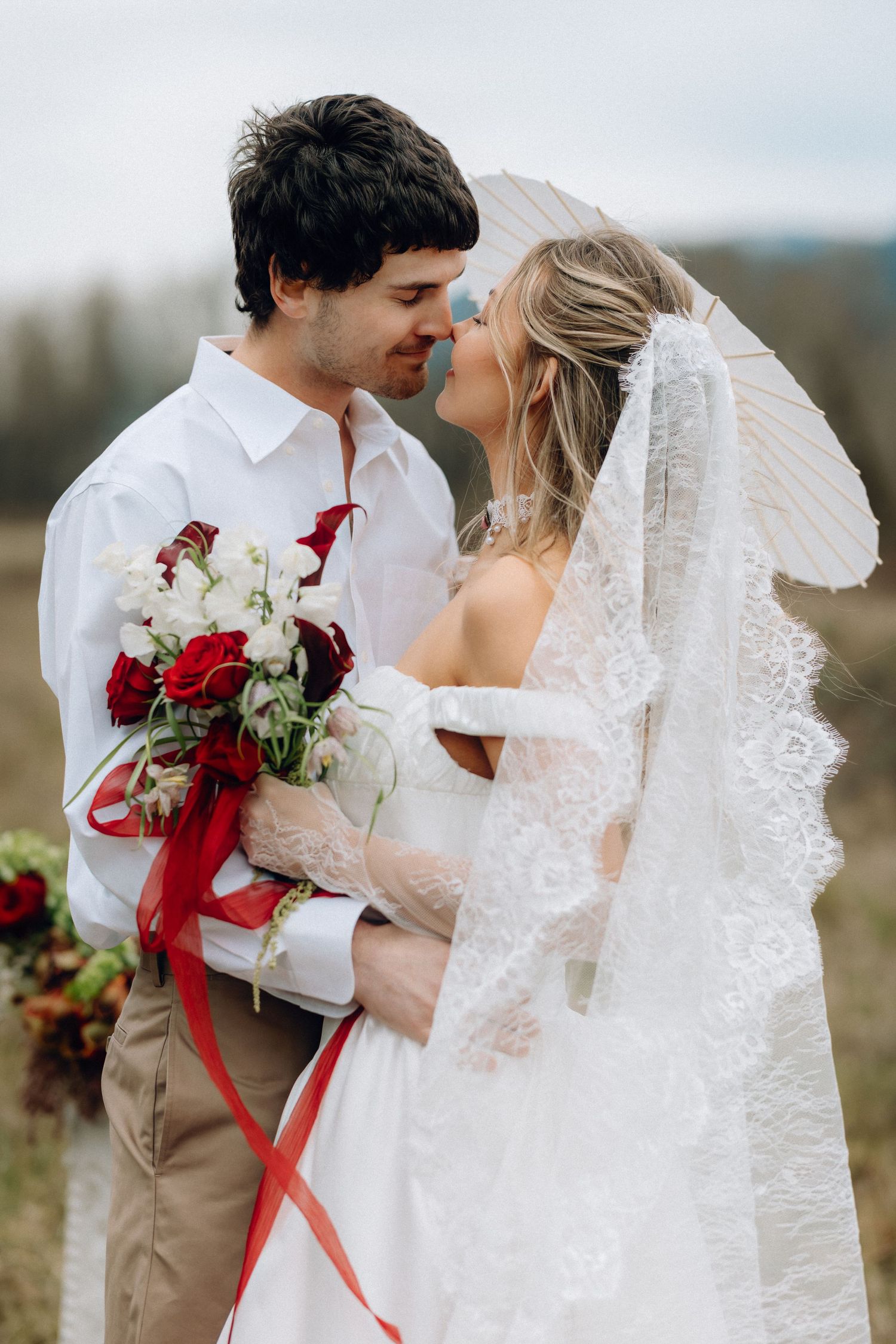 Intimate wedding portraits captured in a field with red bouquet and lace veil details.