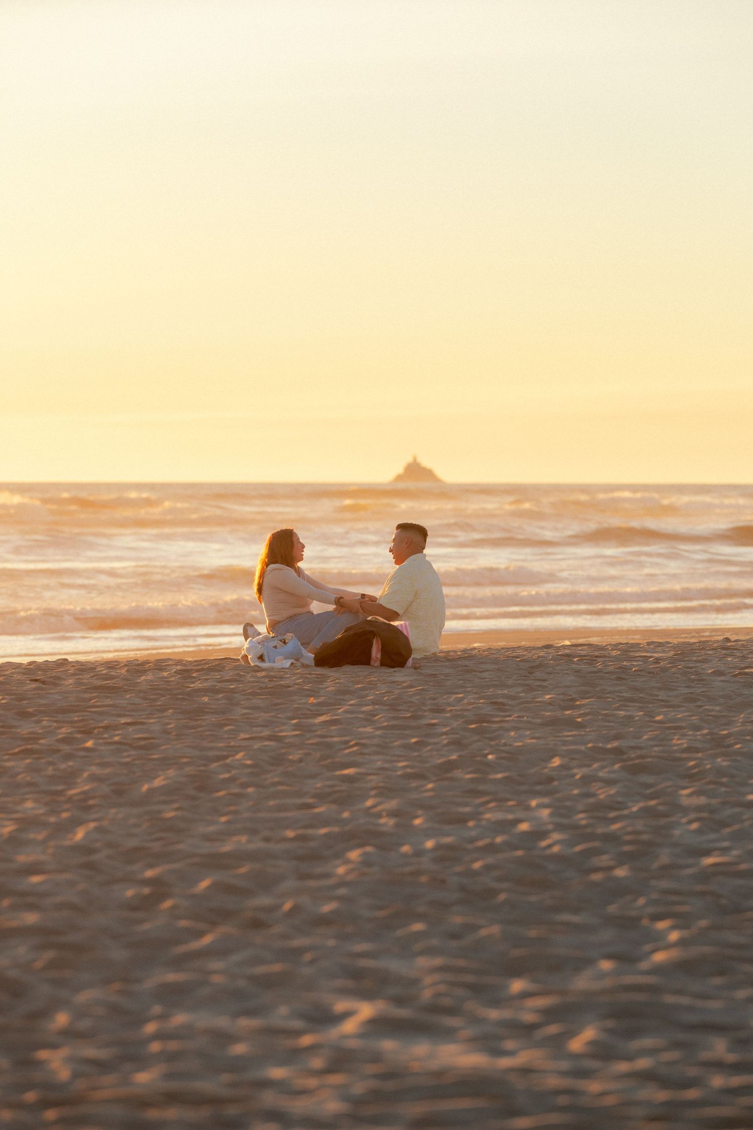 Couple sitting together on a sandy beach at golden sunset with ocean waves and a distant rock formation in the background.