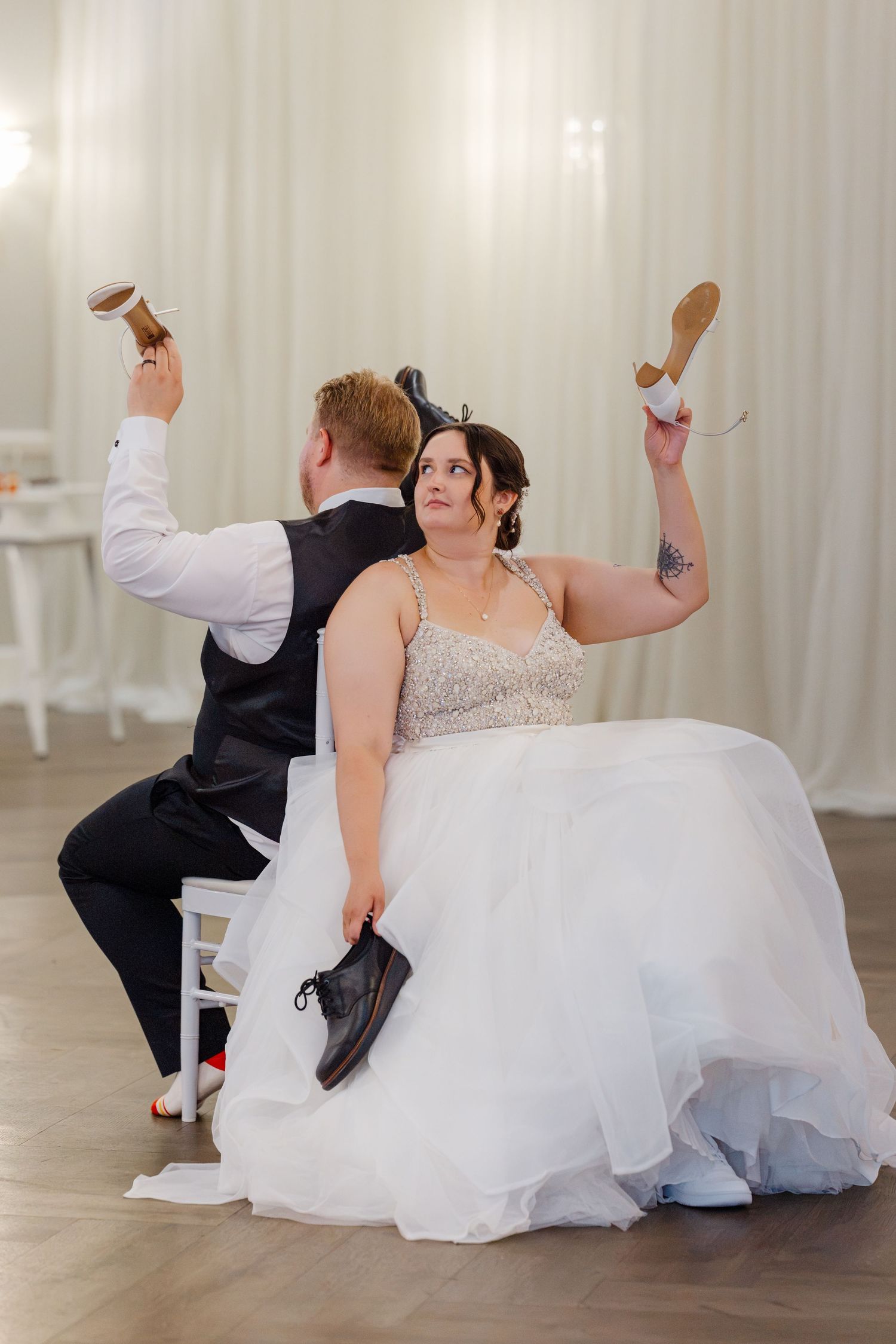Bride and groom play the shoe game at their wedding reception, each holding up a shoe while seated.