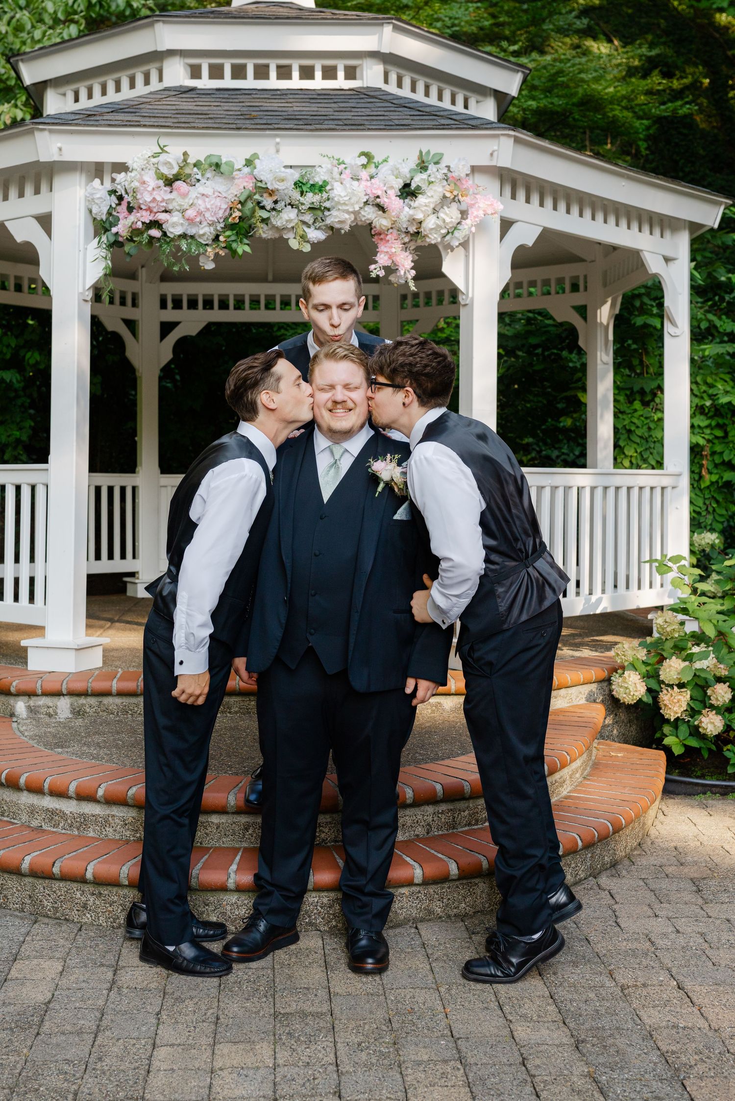 Groom in navy suit being kissed on cheeks by groomsmen in vests before a decorated white gazebo.