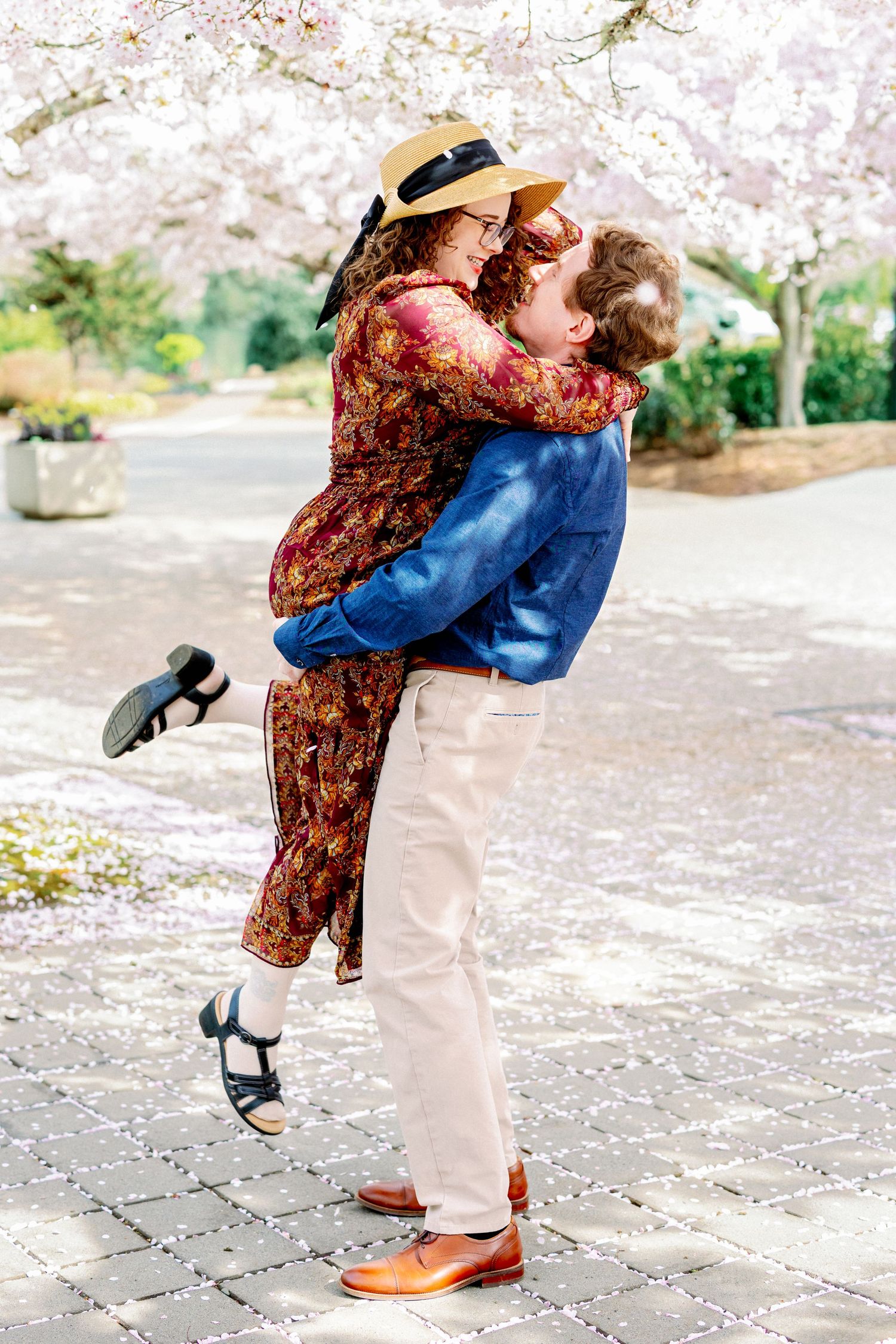 A couple embraces and kisses under blooming cherry blossom trees on a stone pathway in spring.
