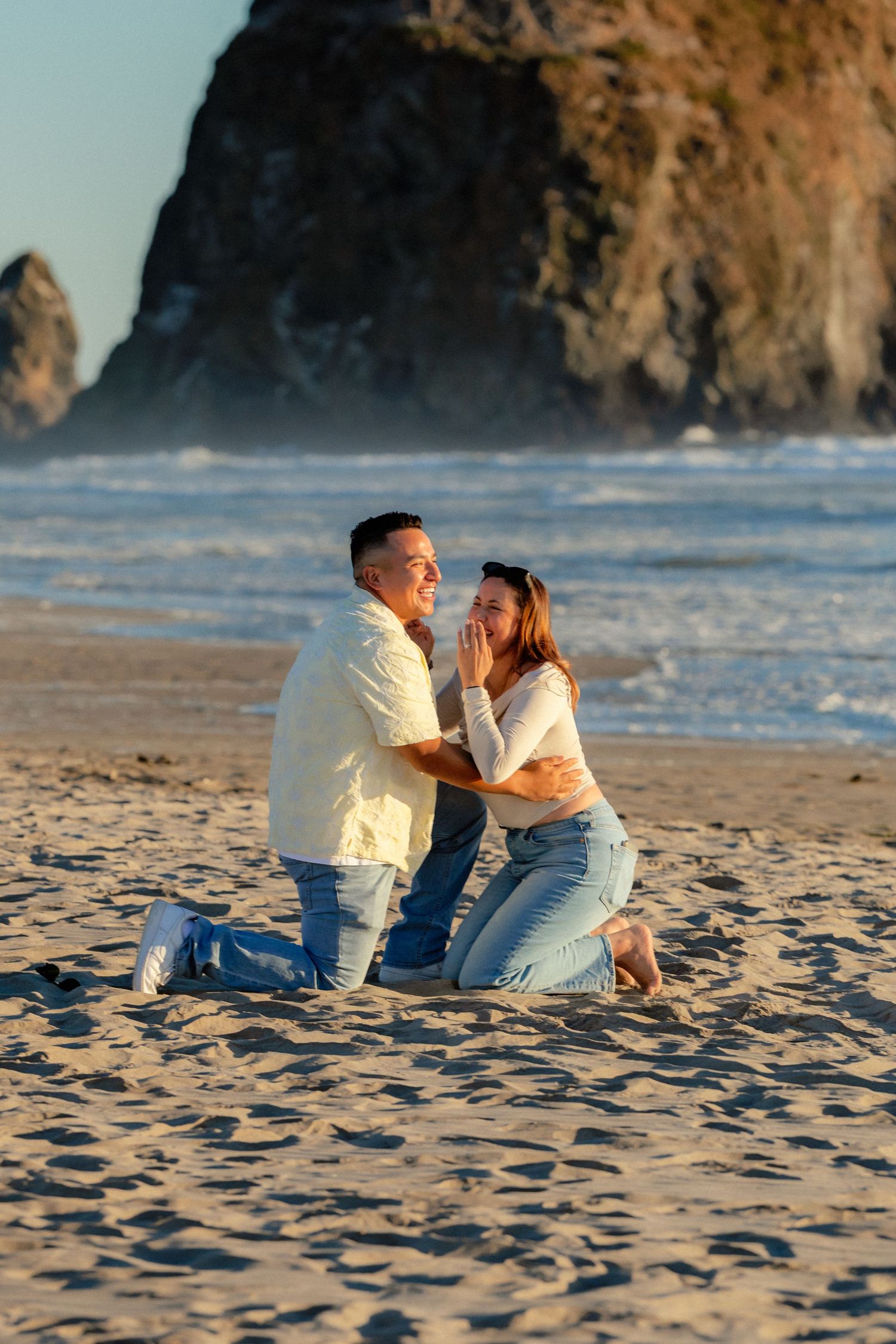 Couple kneeling on sandy beach during golden hour with rocky sea stacks in background, sharing a romantic proposal moment.