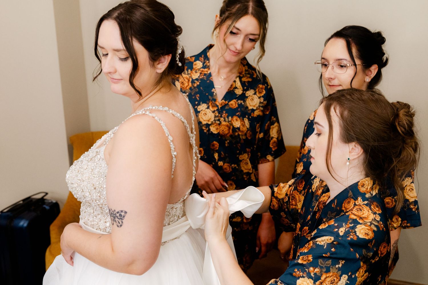 Bridesmaids in floral robes help bride lace up her beaded wedding gown before the ceremony.