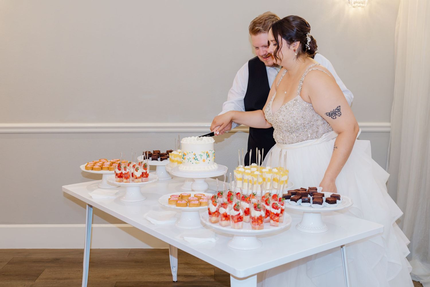 Bride and groom smile while cutting their wedding cake at a dessert table.