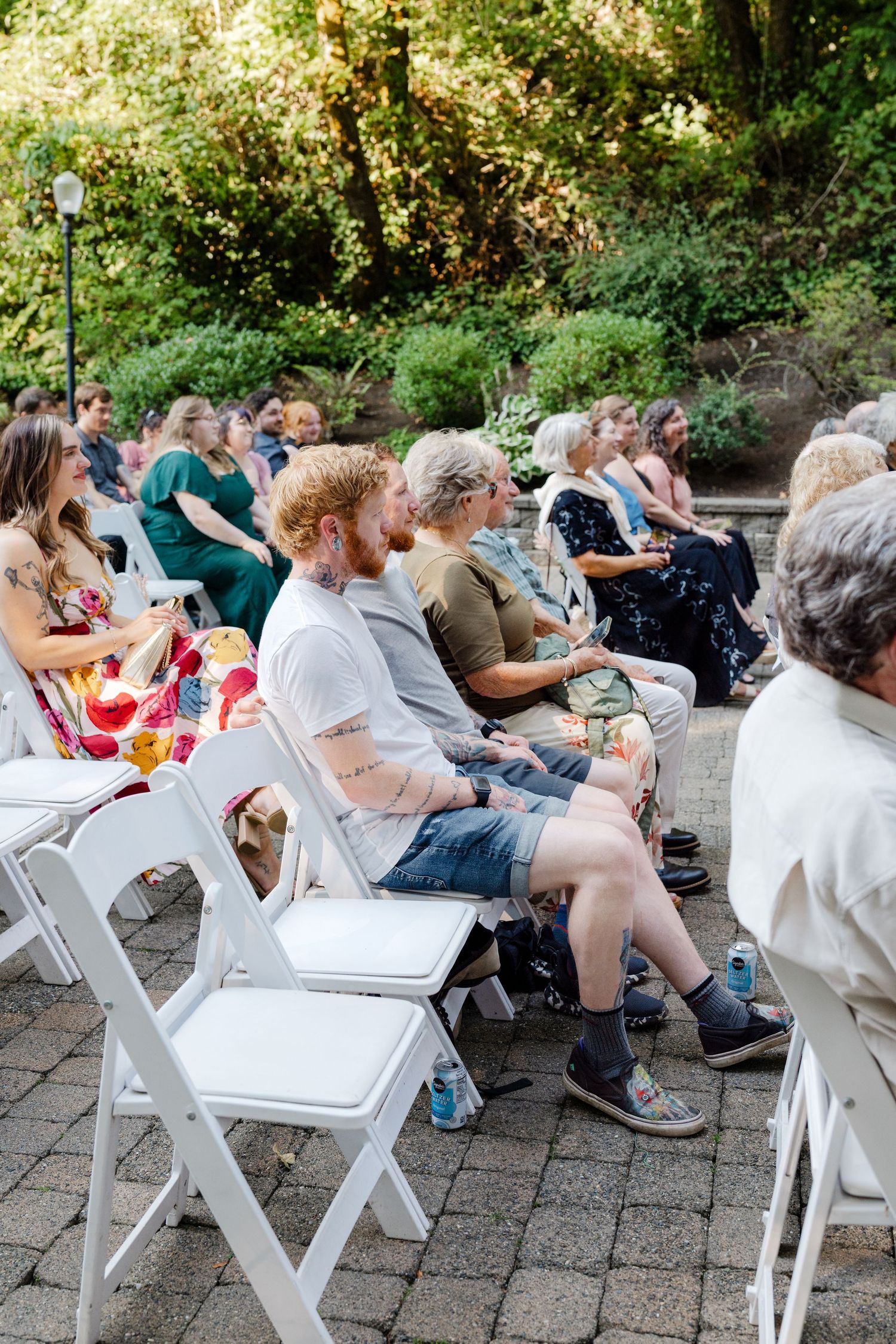 Guests seated in white folding chairs at an outdoor wedding ceremony surrounded by lush greenery.