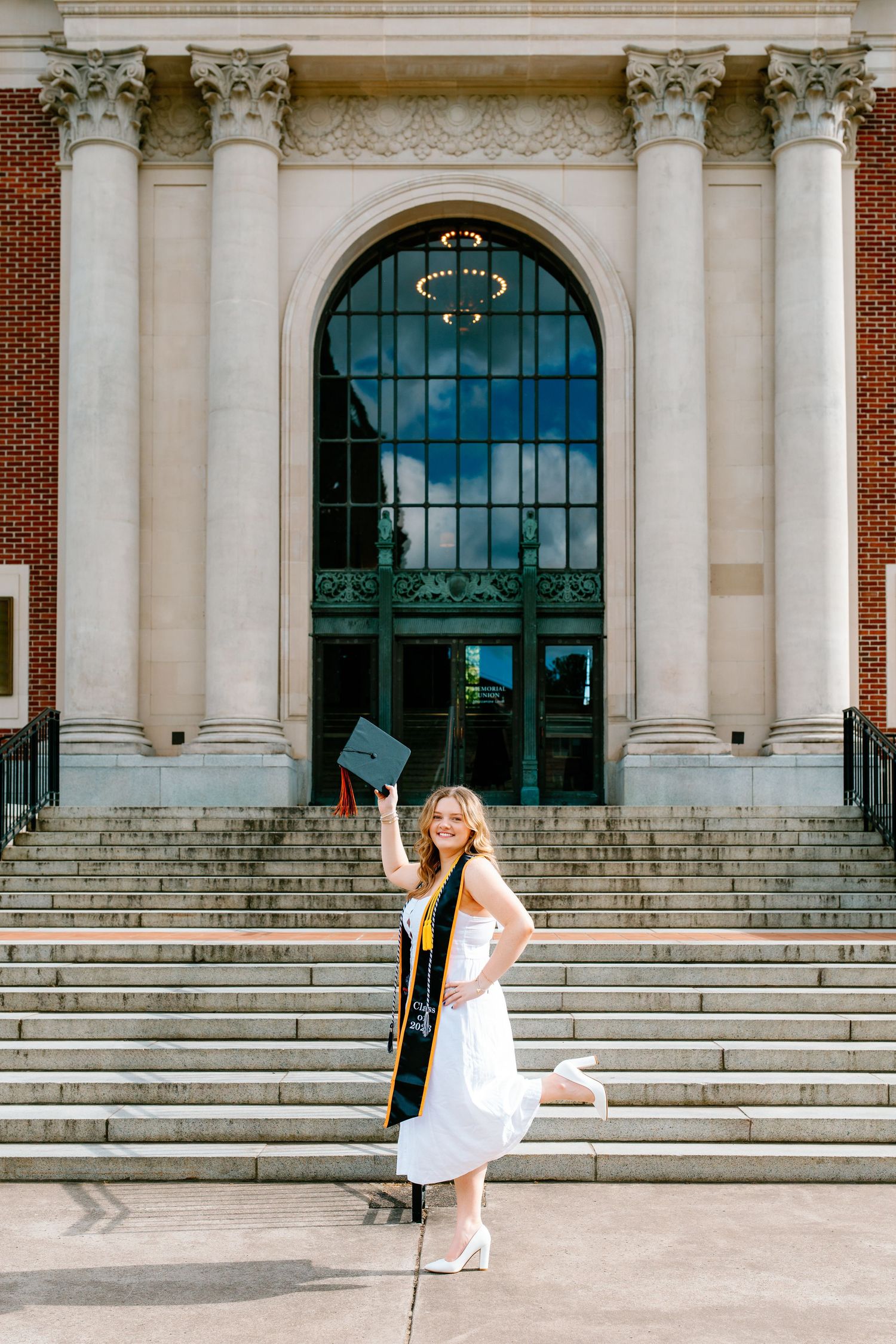 Smiling female graduate in white dress holding cap on steps of brick building with columns.