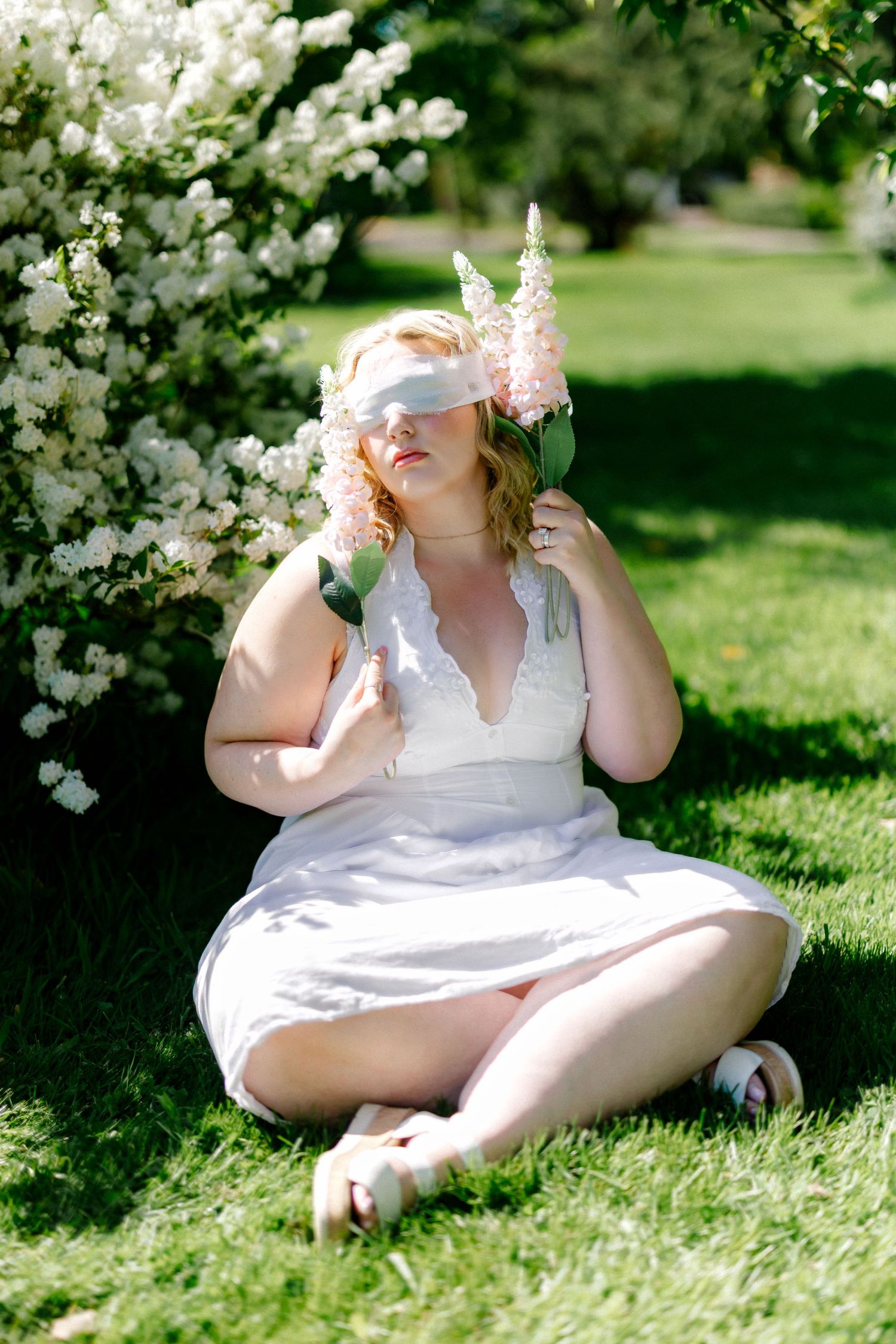 Blonde woman in white dress sits on grass holding pink flowers near white flowering shrubs.