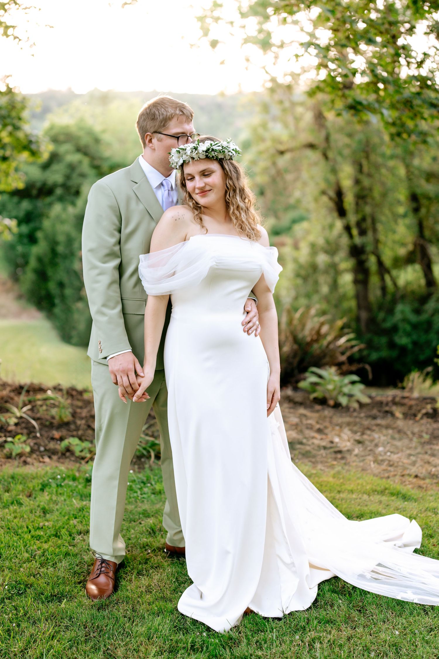 Bride in white off-shoulder gown with floral crown stands with groom in sage suit outdoors.