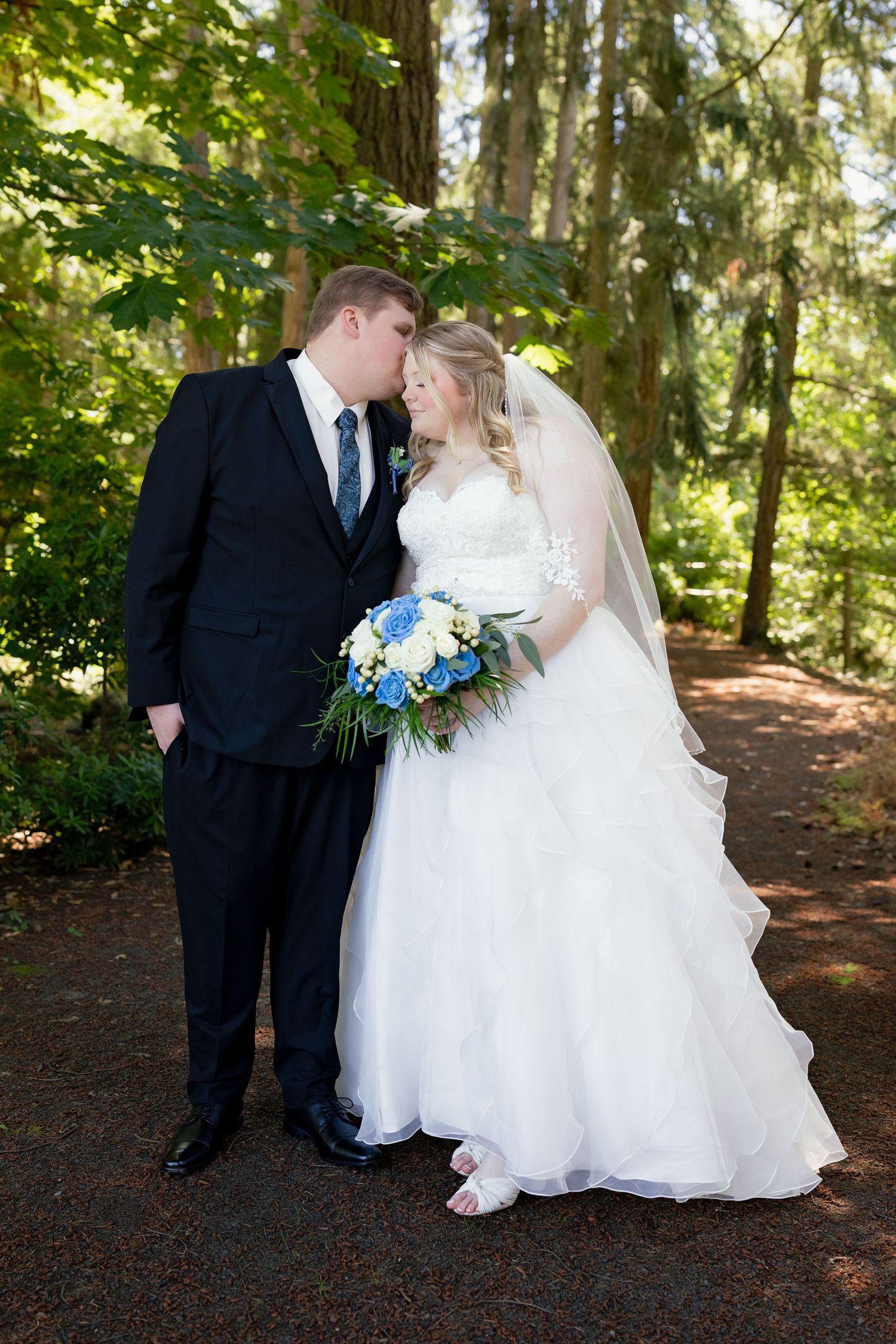 Bride and groom sharing a romantic forehead touch moment on a wooded path, bride holding blue and white bouquet.